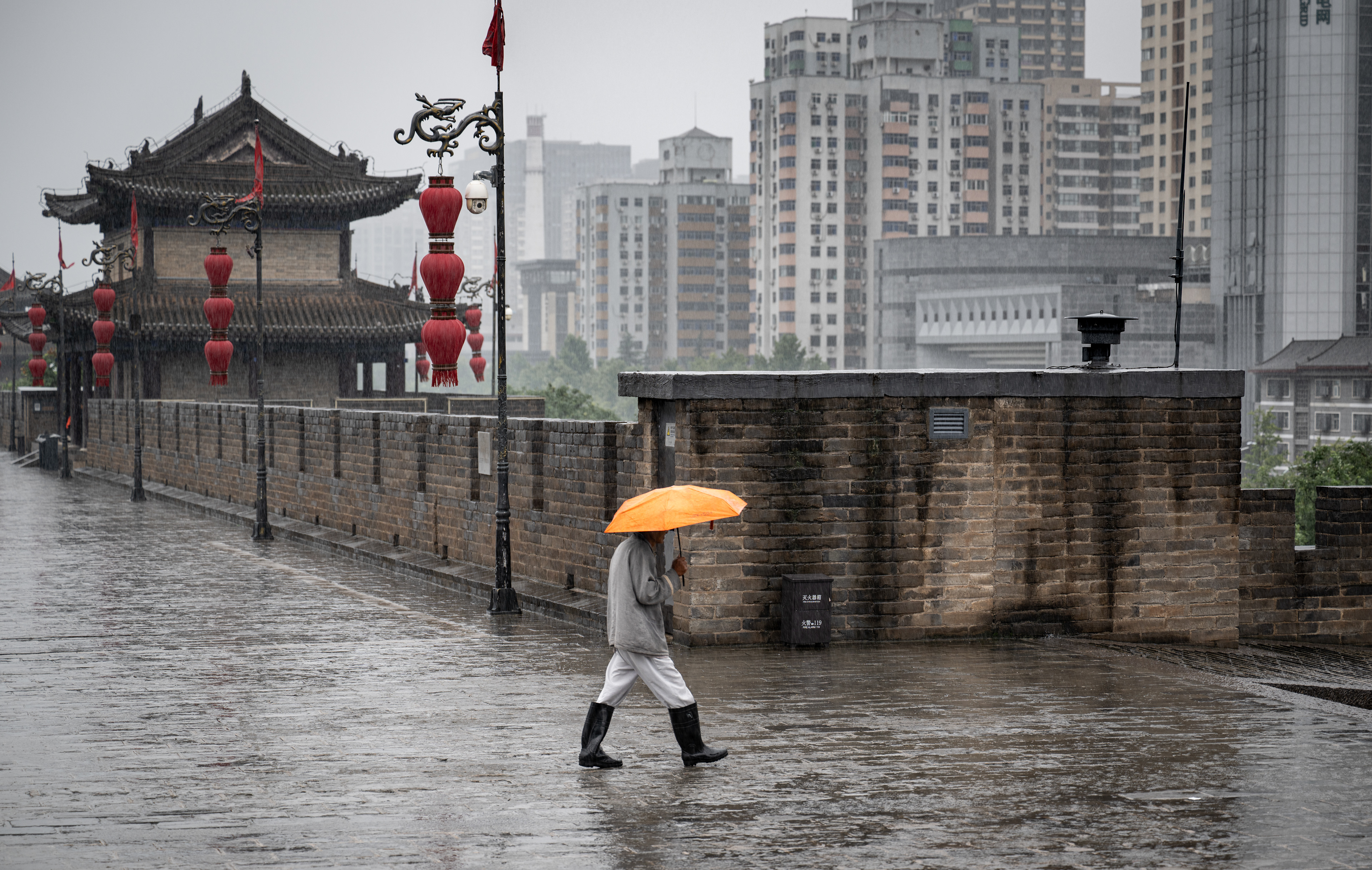 Sulle mura di Xi'an, sotto la pioggia, un uomo con un ombrello arancione cammina verso i grattacieli della città nuova — Fabio Alimonti