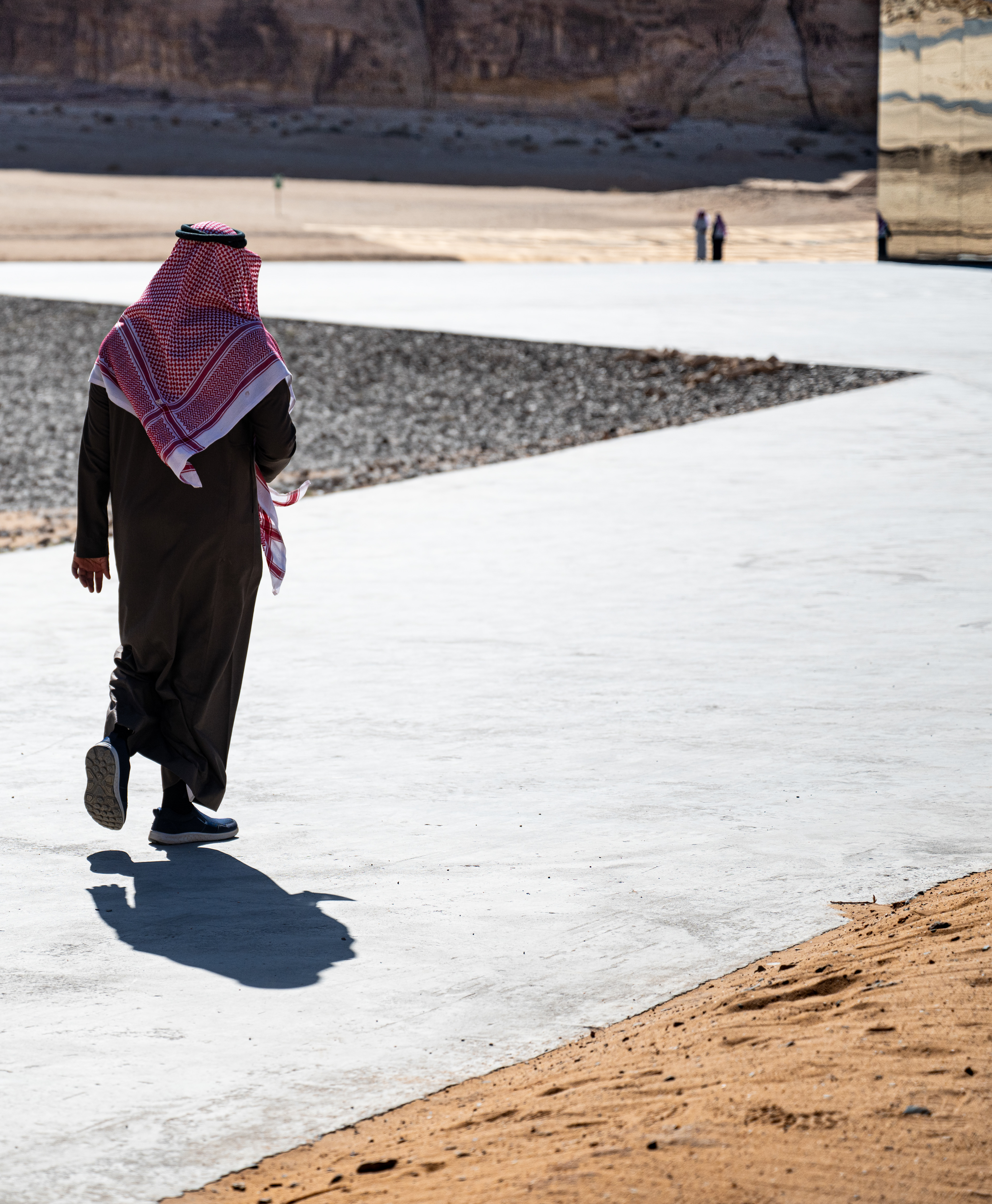 Appuntamento al Maraya – un uomo in thobe e kefiah rossa cammina su un sentiero di cemento ad AlUla verso due figure lontane, accanto all'edificio a specchio Maraya. Fotografia di Fabio Alimonti.