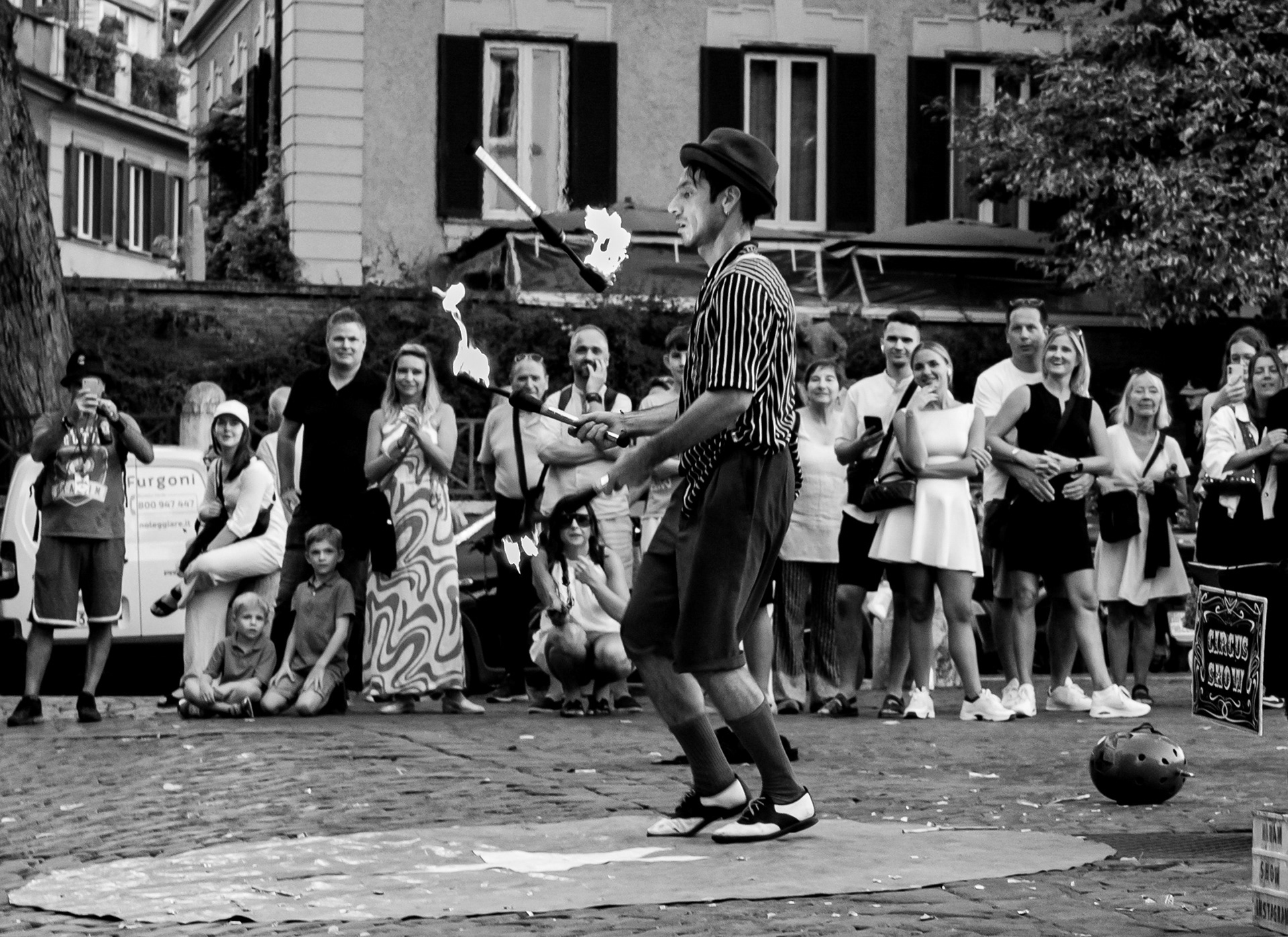 Circus Show – bianco e nero: un giocoliere con bombetta e camicia a righe fa roteare clave infuocate in una piazza di Trastevere, davanti a un pubblico che guarda e riprende con i telefoni. Fotografia di Fabio Alimonti.