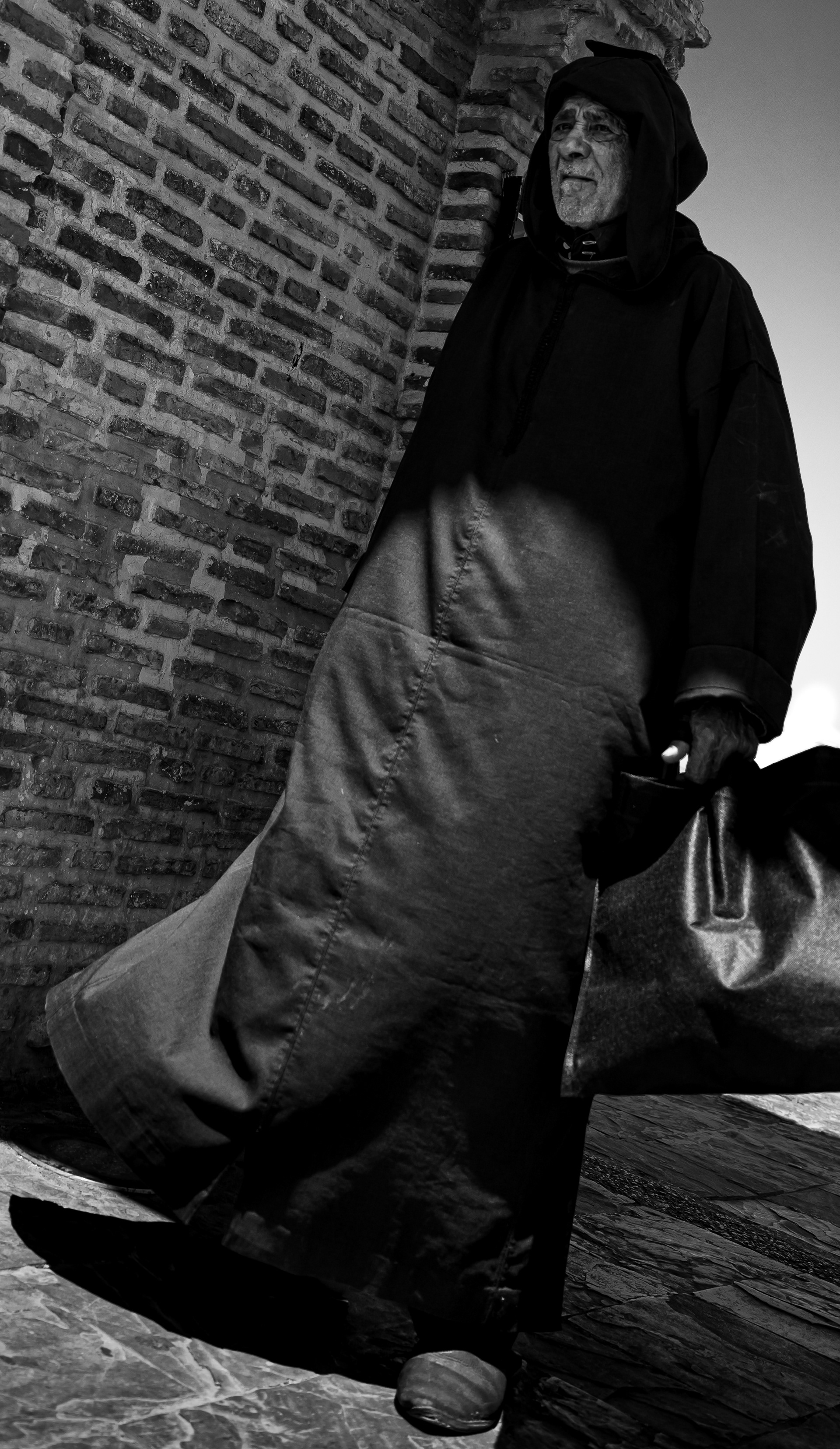 Il custode si è alzato – anziano in djellaba con cappuccio che cammina lungo un muro di mattoni, metà nella luce e metà nell'ombra, Tangeri, Marocco. Fotografia in bianco e nero di Fabio Alimonti