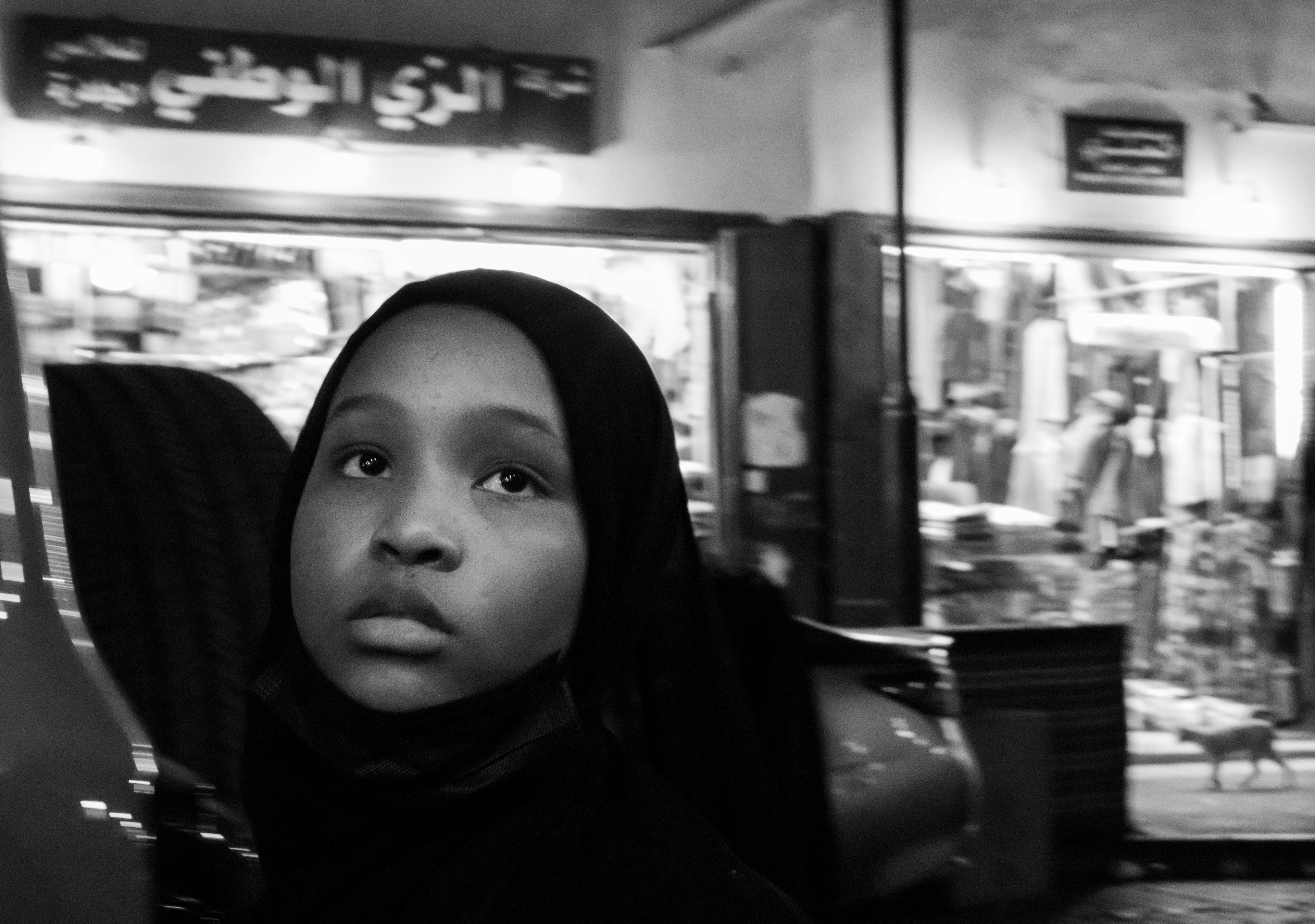 Dove la luce si ferma: ritratto in bianco e nero di una bambina con hijab che guarda in alto, vetrine del suq di Al-Balad a Gedda sullo sfondo, fotografia di Fabio Alimonti