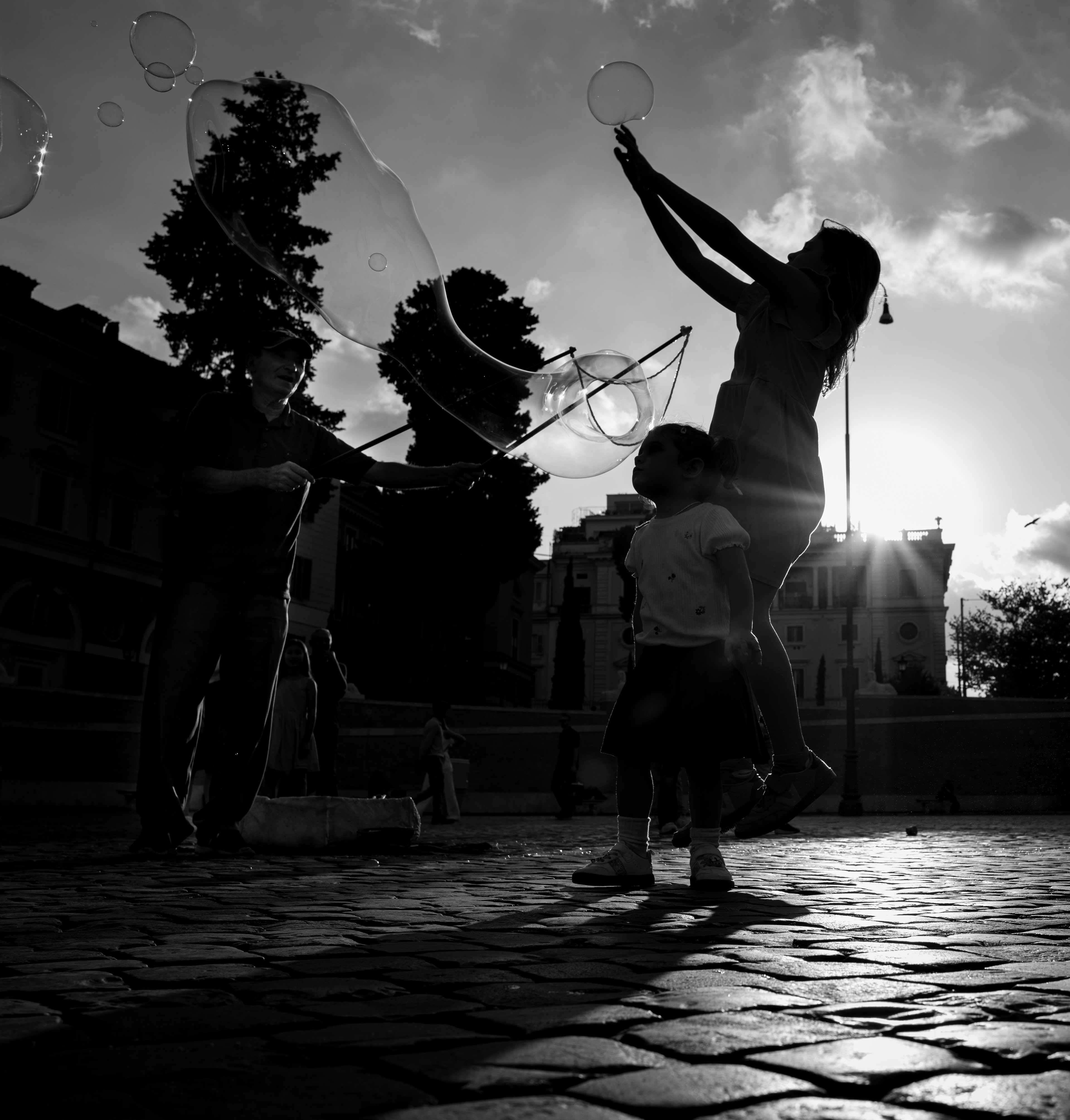 Quasi – in Piazza del Popolo un uomo fa una bolla di sapone gigante, una bambina in controluce salta per prenderla e una più piccola la guarda, Roma. Fotografia in bianco e nero di Fabio Alimonti