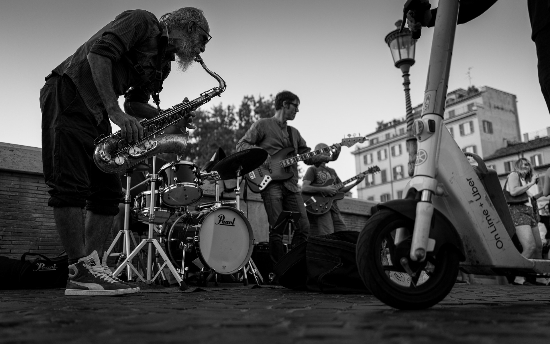 Controtempo – bianco e nero: un sassofonista anziano con la barba bianca si piega sul suo strumento mentre la band suona su Ponte Sisto; in primo piano un monopattino a noleggio, sullo sfondo una ragazza che balla. Fotografia di Fabio Alimonti.