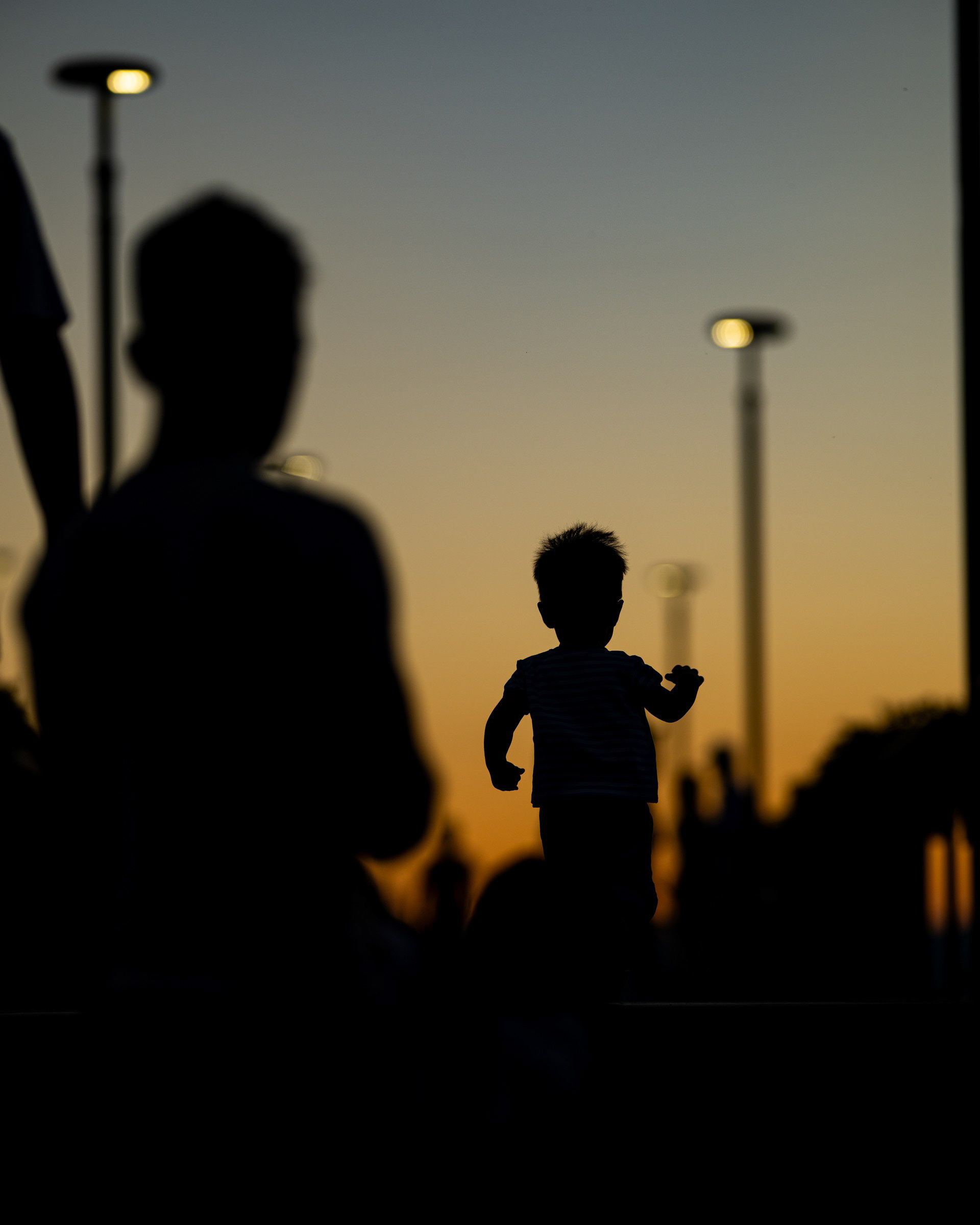 Un bambino corre in controluce sul lungolago di Trevignano Romano al tramonto, davanti alla sagoma sfocata del padre che lo osserva
