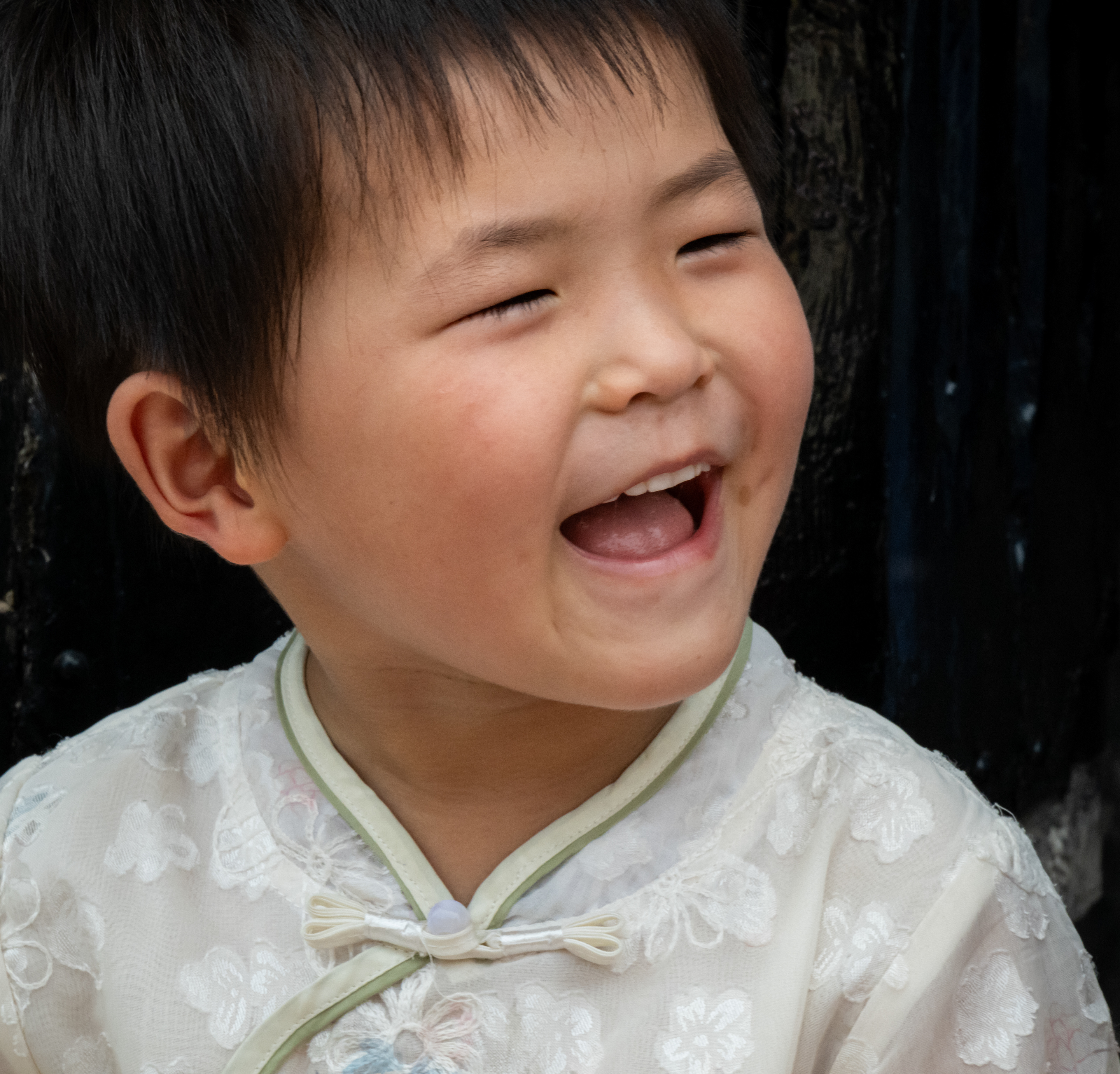 Bambino che ride con gli occhi chiusi, giacca bianca ricamata contro un muro nero, Pingyao, Cina
