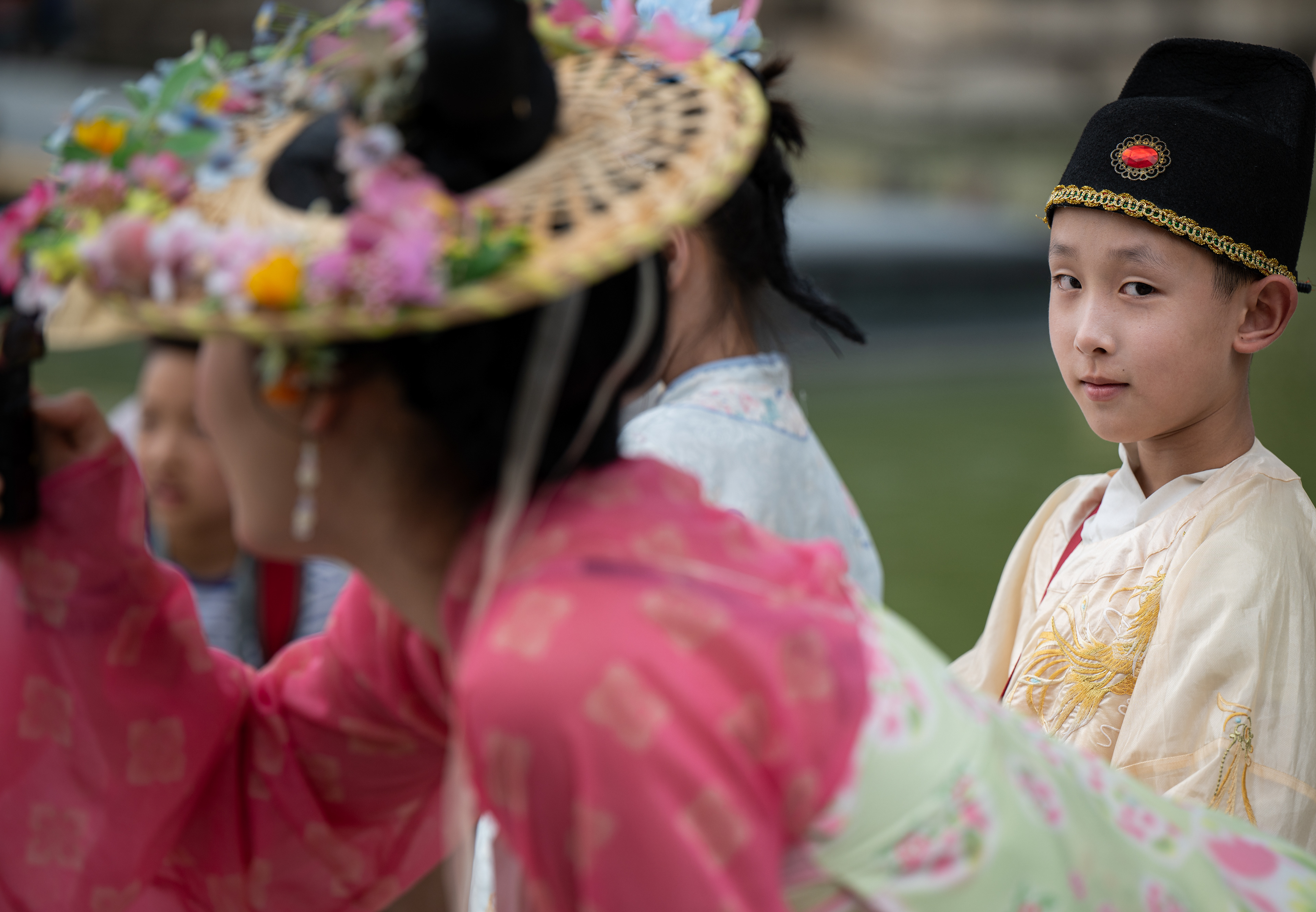 Bambino in veste gialla e cappello nero da funzionario guarda in macchina, davanti una donna in abito rosa con cappello fiorito fotografa la pagoda, Mutianyu, Cina