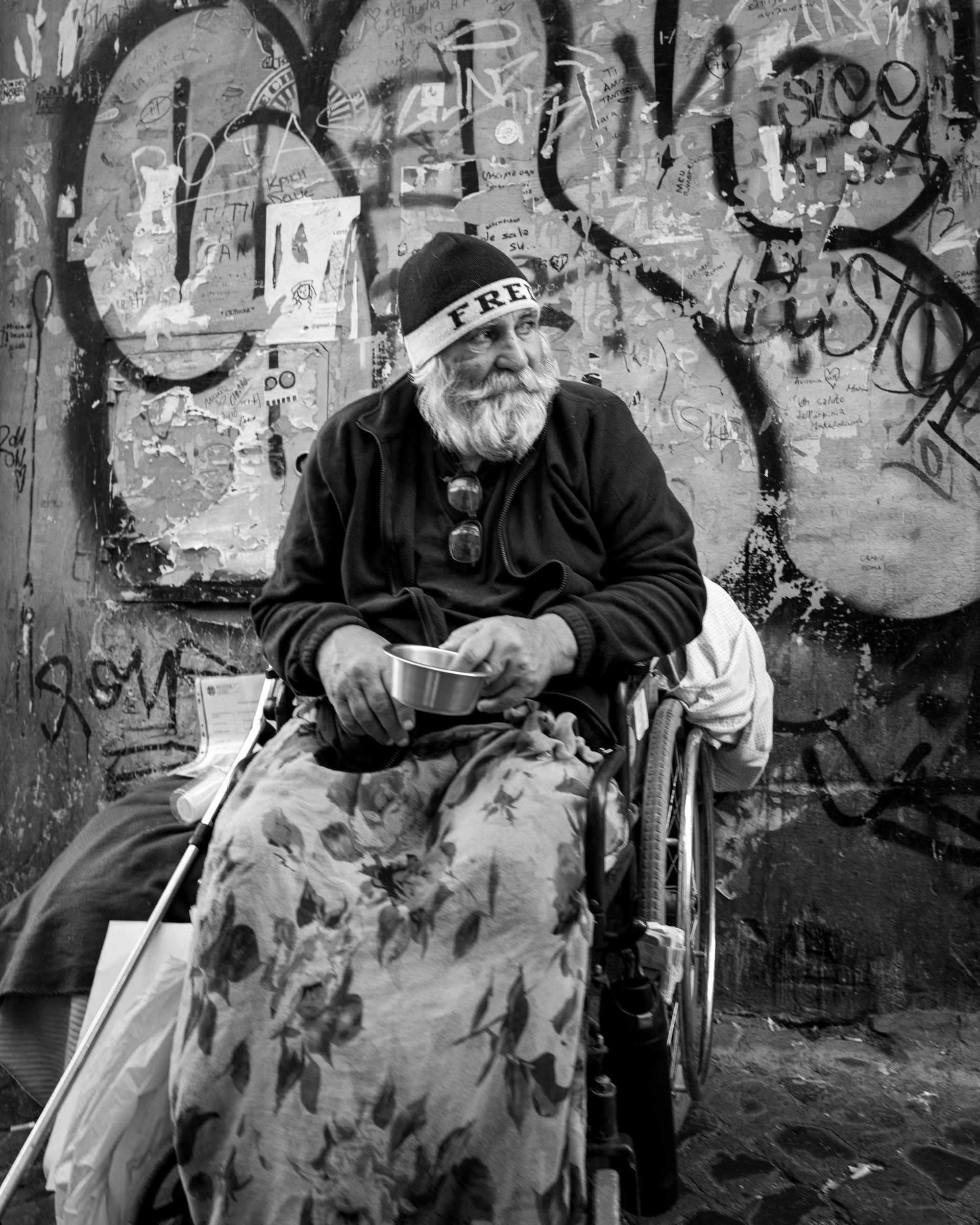 Free – ritratto in bianco e nero di un uomo anziano con la barba bianca e un berretto con la scritta FREE, davanti a un muro coperto di graffiti a Trastevere, Roma. Fotografia di Fabio Alimonti.