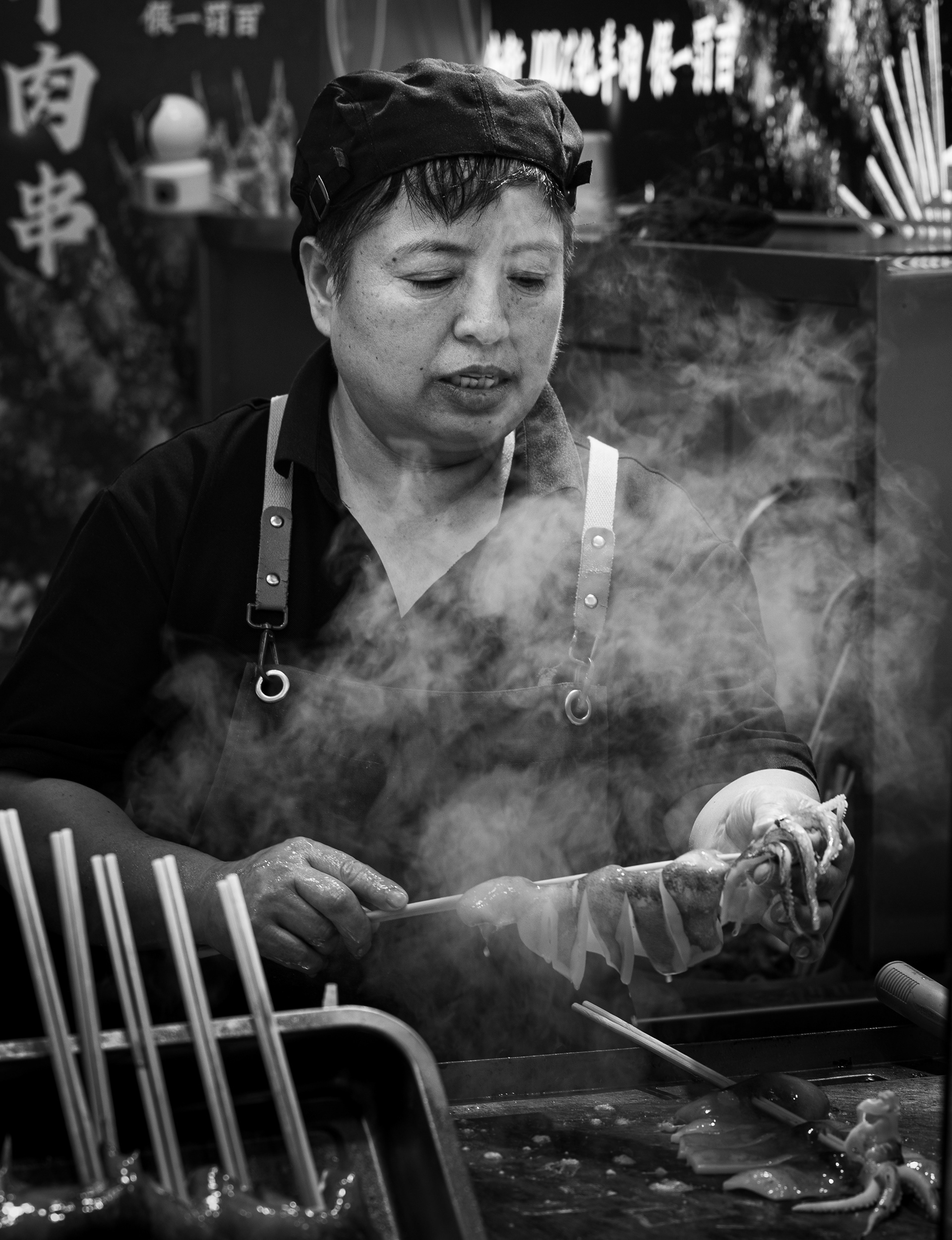 Nel vapore – fotografia in bianco e nero di una cuoca a un banco di spiedini a Datong, Cina, che infila un polpo su uno spiedo tra il vapore