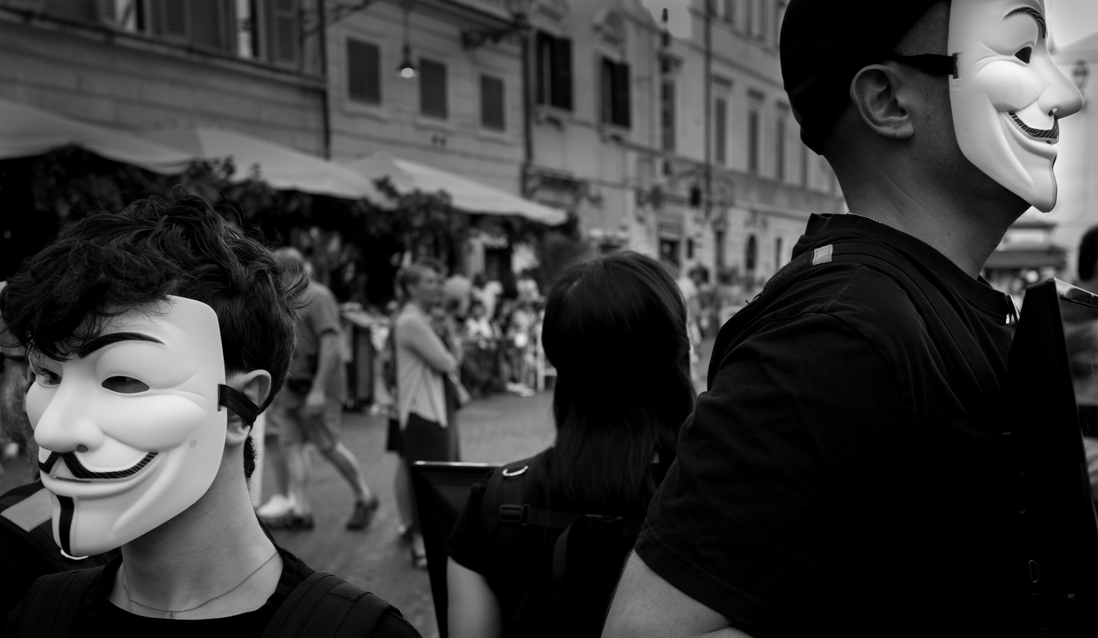 Due persone con la maschera di Anonymous tra la folla in una piazza di Roma, in bianco e nero