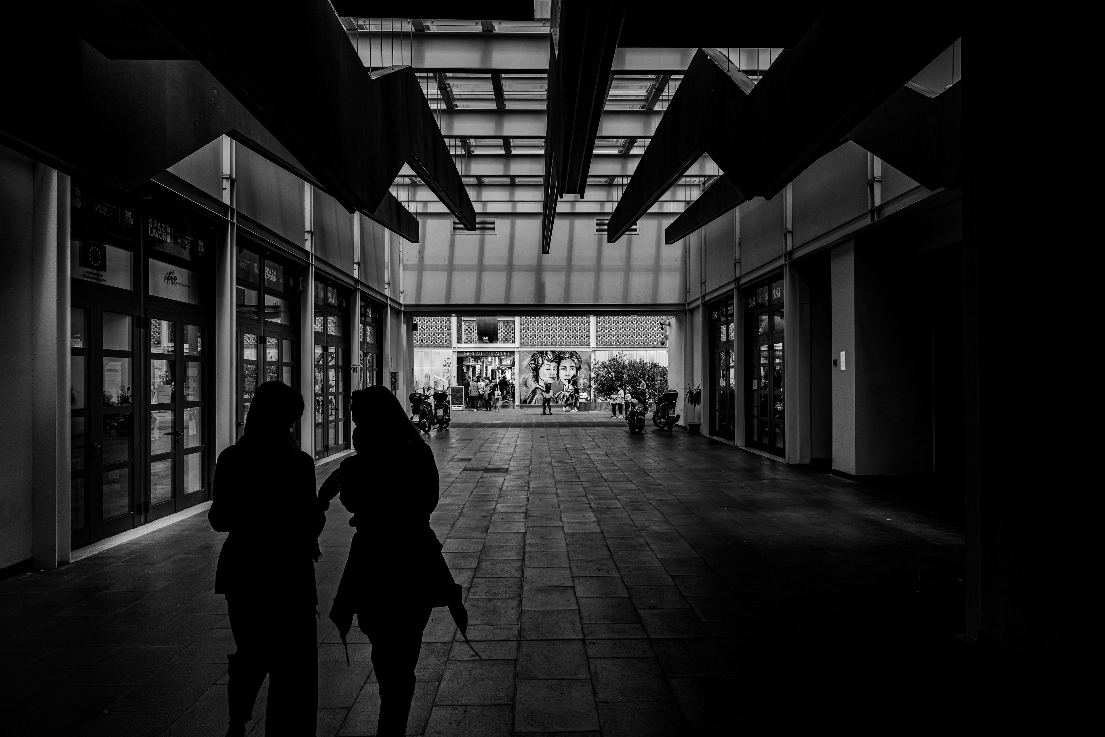Verso la luce, tra ombre sospese – fotografia in bianco e nero di Fabio Alimonti: due sagome camminano verso l'apertura luminosa di un mercato coperto a Roma