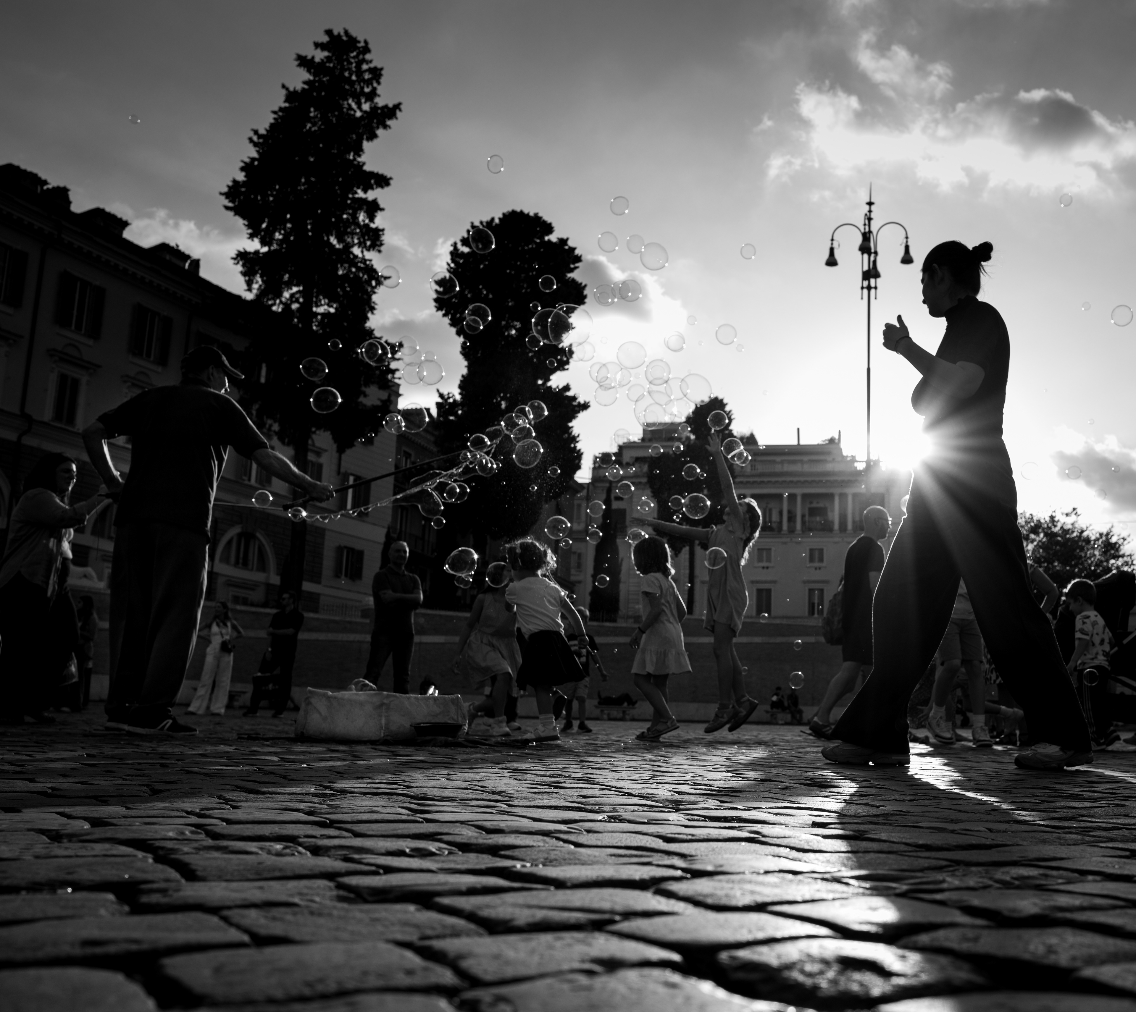 Un respiro di luce – in Piazza del Popolo un uomo fa le bolle di sapone, i bambini le rincorrono e una donna in controluce attraversa il sole, Roma. Fotografia in bianco e nero di Fabio Alimonti