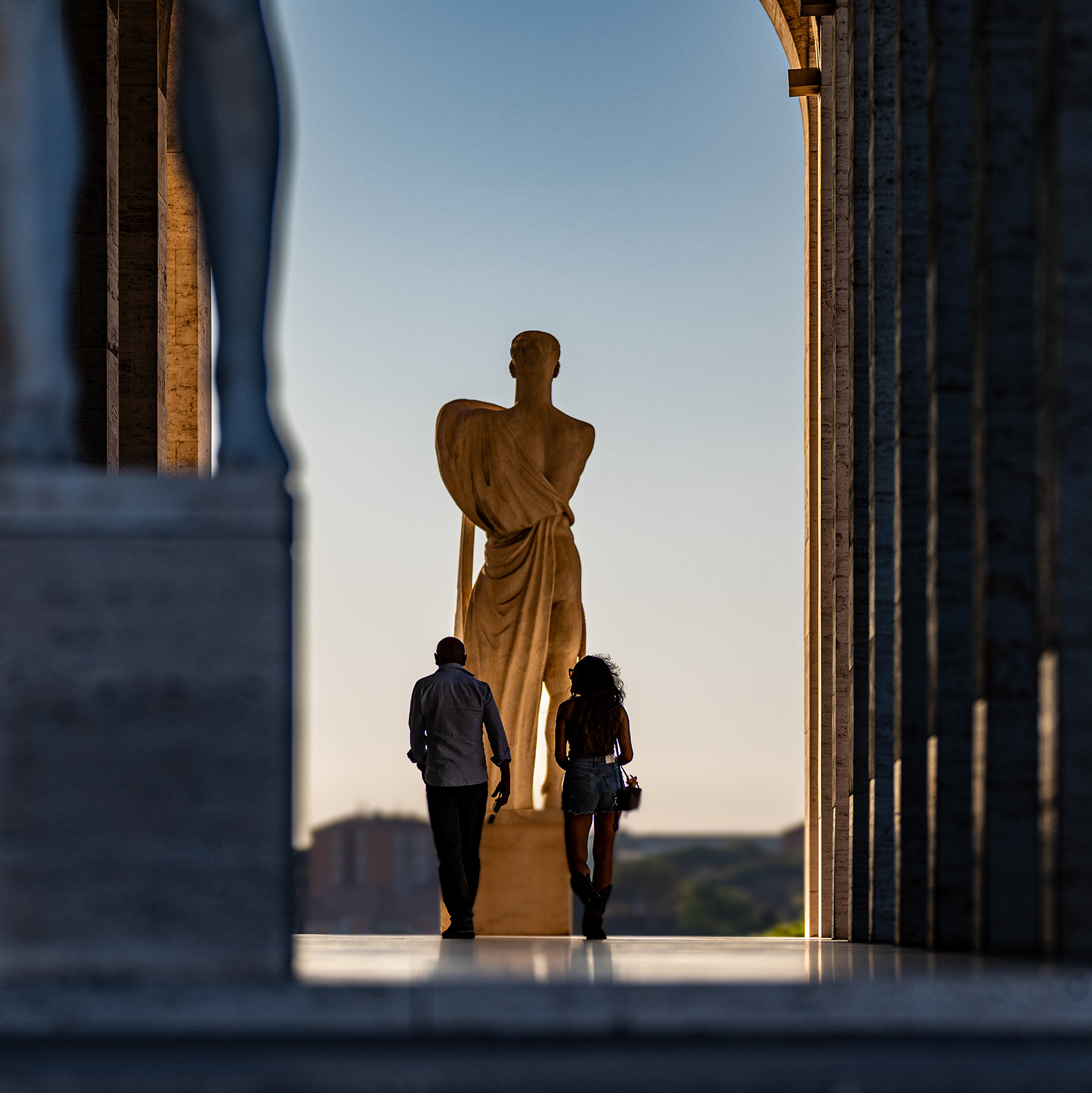 Ombre di passaggio – una coppia cammina in controluce sotto la statua di marmo nell'arco del Palazzo della Civiltà Italiana, EUR, Roma. Foto di Fabio Alimonti