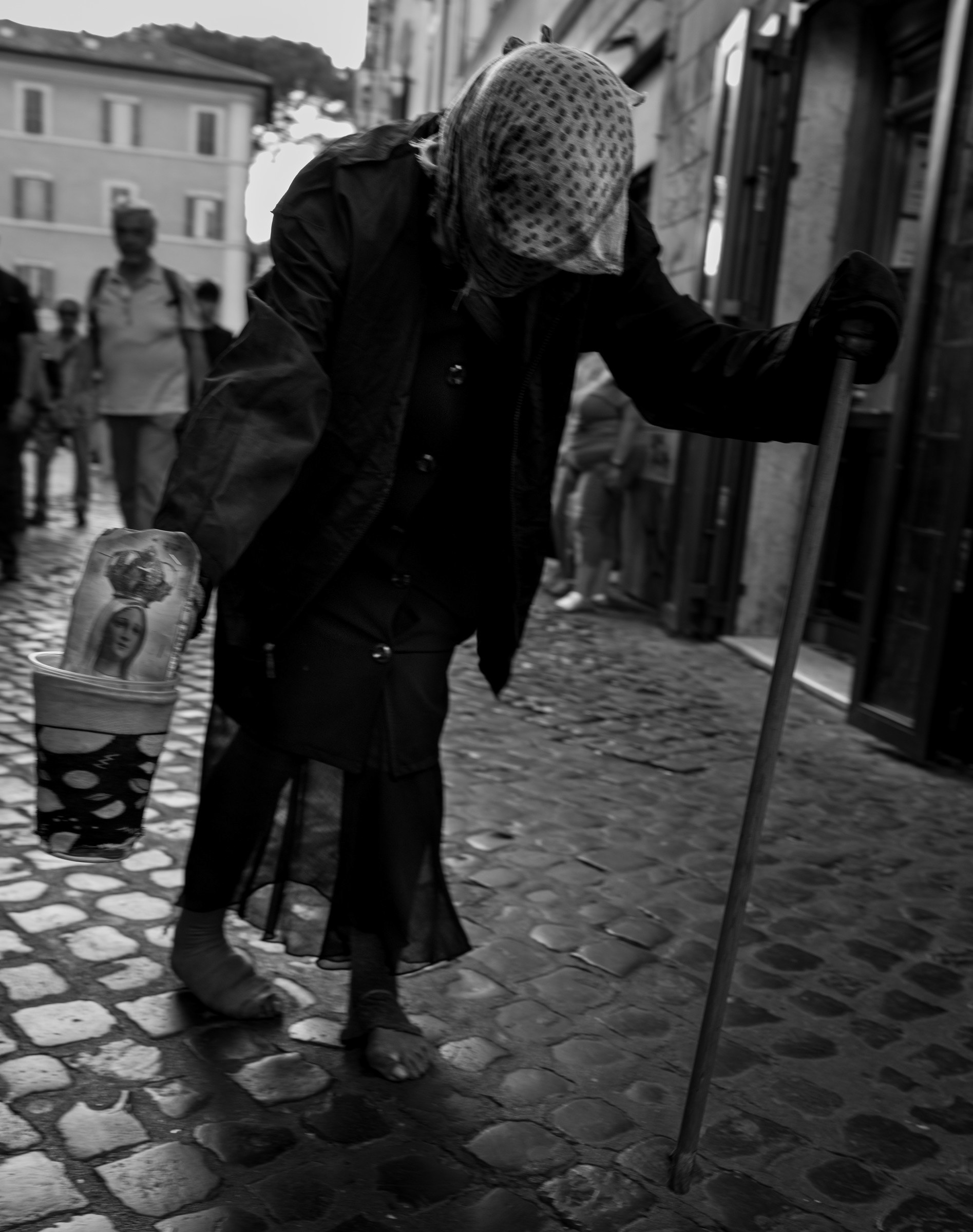 In compagnia – bianco e nero: un'anziana con il capo coperto da un fazzoletto a pois, curva sul bastone, cammina sui sampietrini di Trastevere portando un bicchiere con l'immagine della Madonna. Fotografia di Fabio Alimonti.