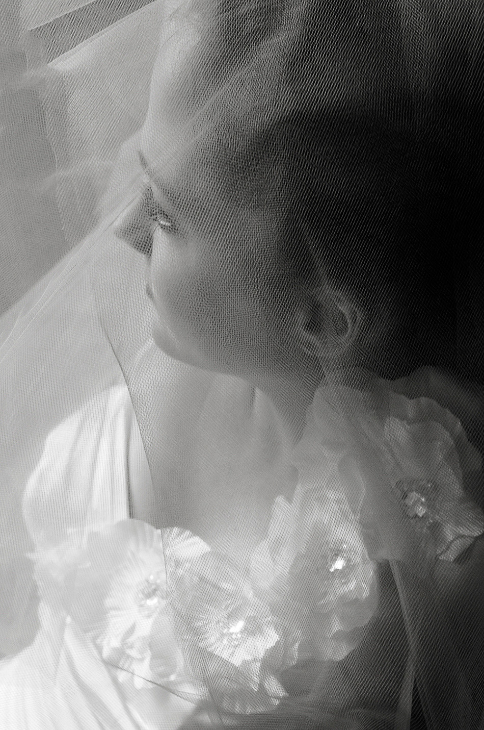 A close-up black and white portrait of a bride in head and shoulders view, her face turned away from the camera, partially veiled in soft lace.