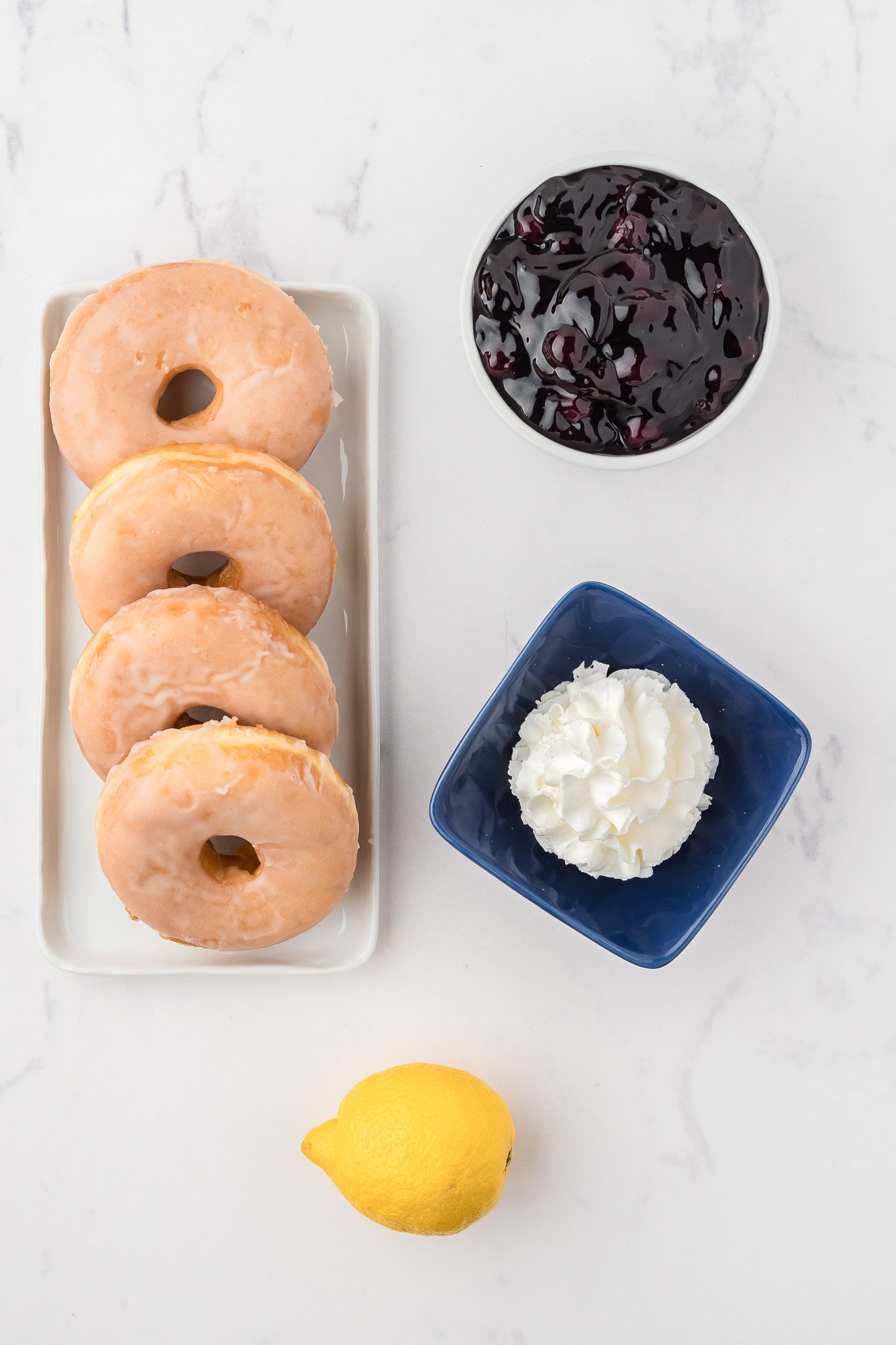 Overhead view of ingredients for blueberry donut shortcakes: glazed donuts, blueberry pie filling, whipped cream, and a lemon on a white marble surface.
