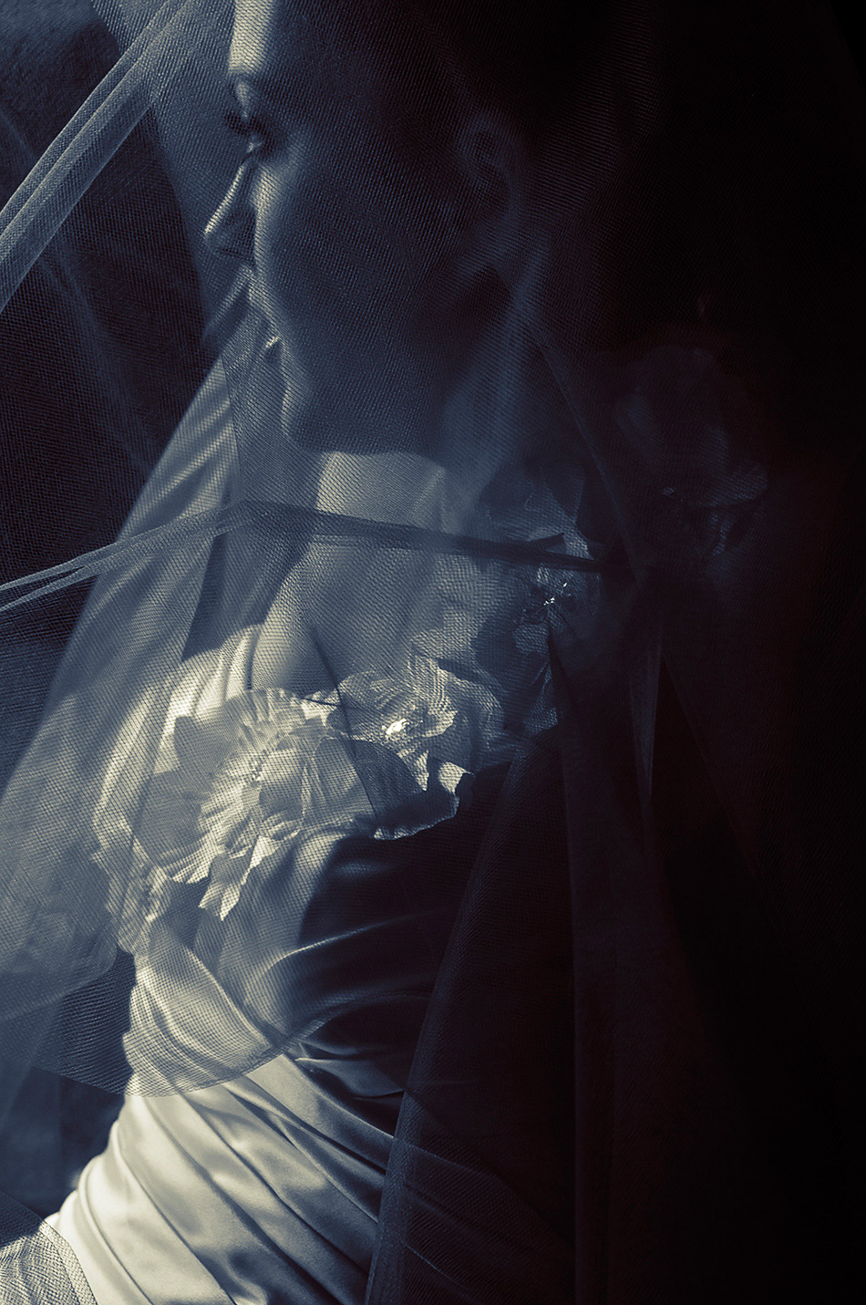 A close-up, blue-toned bridal portrait with deep shadows and a pensive expression, the bride’s eyes cast downward and partially hidden by a veil.