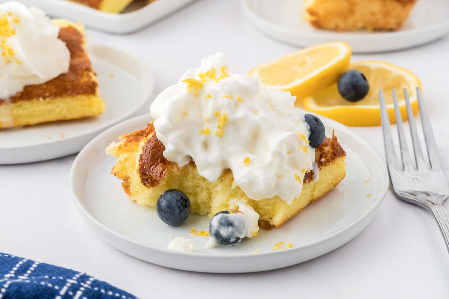 Close-up of a slice of lemon angel food sponge cake topped with whipped cream, lemon zest, and blueberries on a white plate.