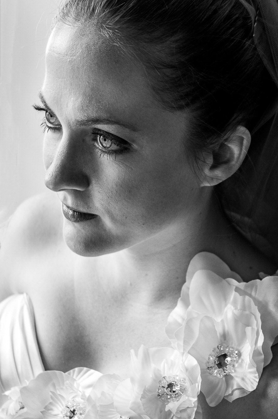 A high-contrast black and white portrait of a bride in head and shoulders view, gazing slightly past the camera with no veil and dramatic lighting across her face.