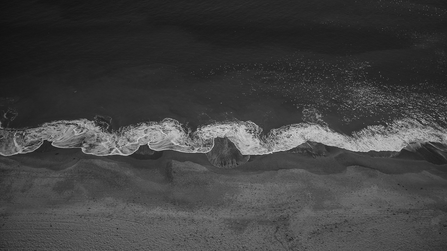 A high-contrast black and white photograph of the shoreline at Myrtle Beach, South Carolina. Captured from directly overhead, the image features a sharply defined line where ocean water meets sand, creating a bold, abstract composition of texture and tension.