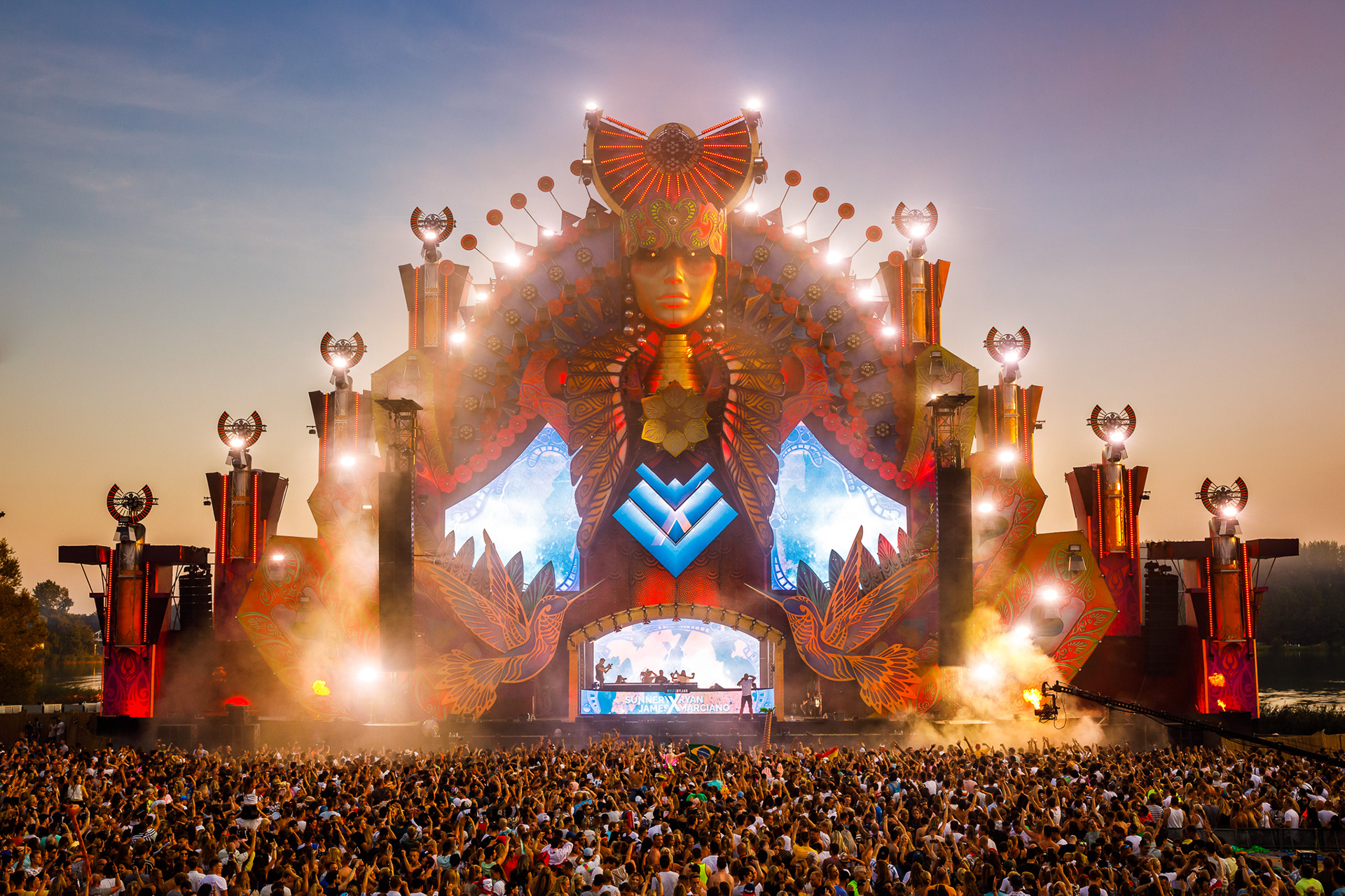 Crowd at a music festival with a large, ornate stage featuring lights and LED screens.