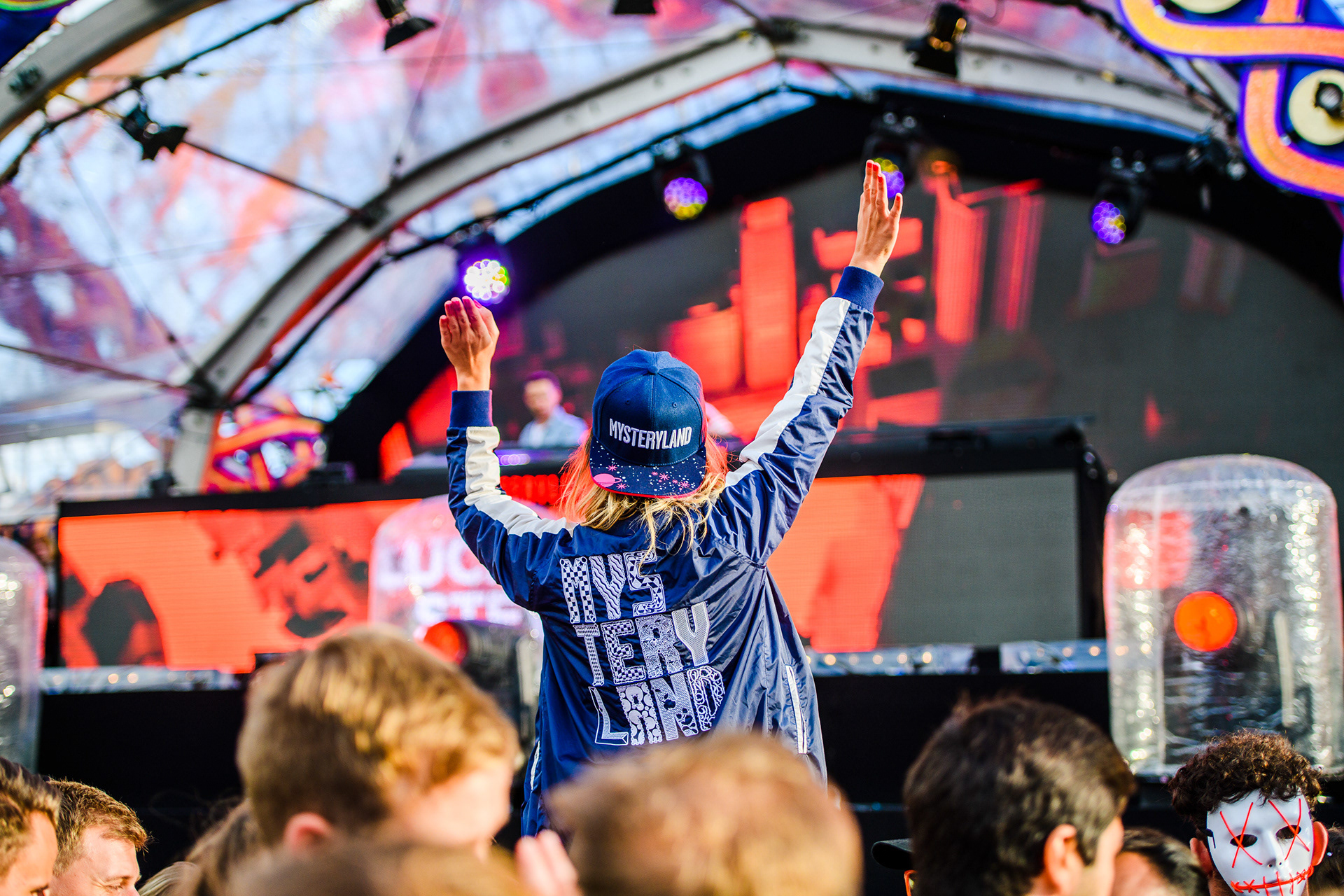 Person at a festival wearing a "Mysteryland" cap, hands raised, with stage in the background.