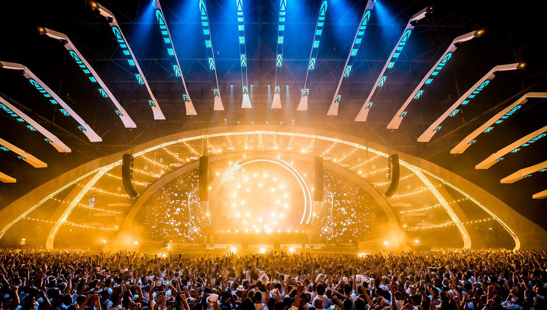 A indoor music event with a large crowd, bright lights, and a vibrant stage setup.