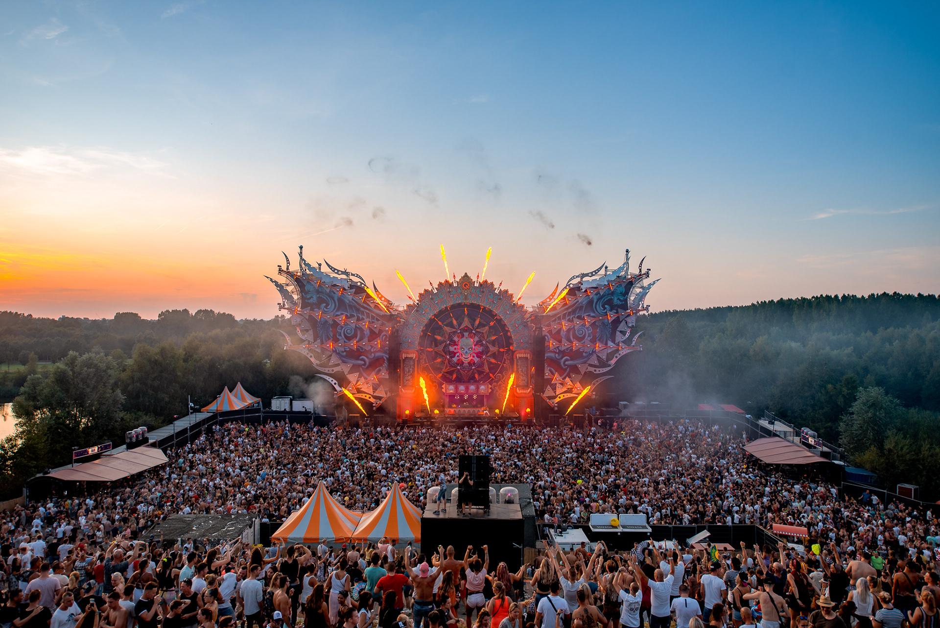 Outdoor music festival crowd with a large, elaborate stage and fireworks at dusk.