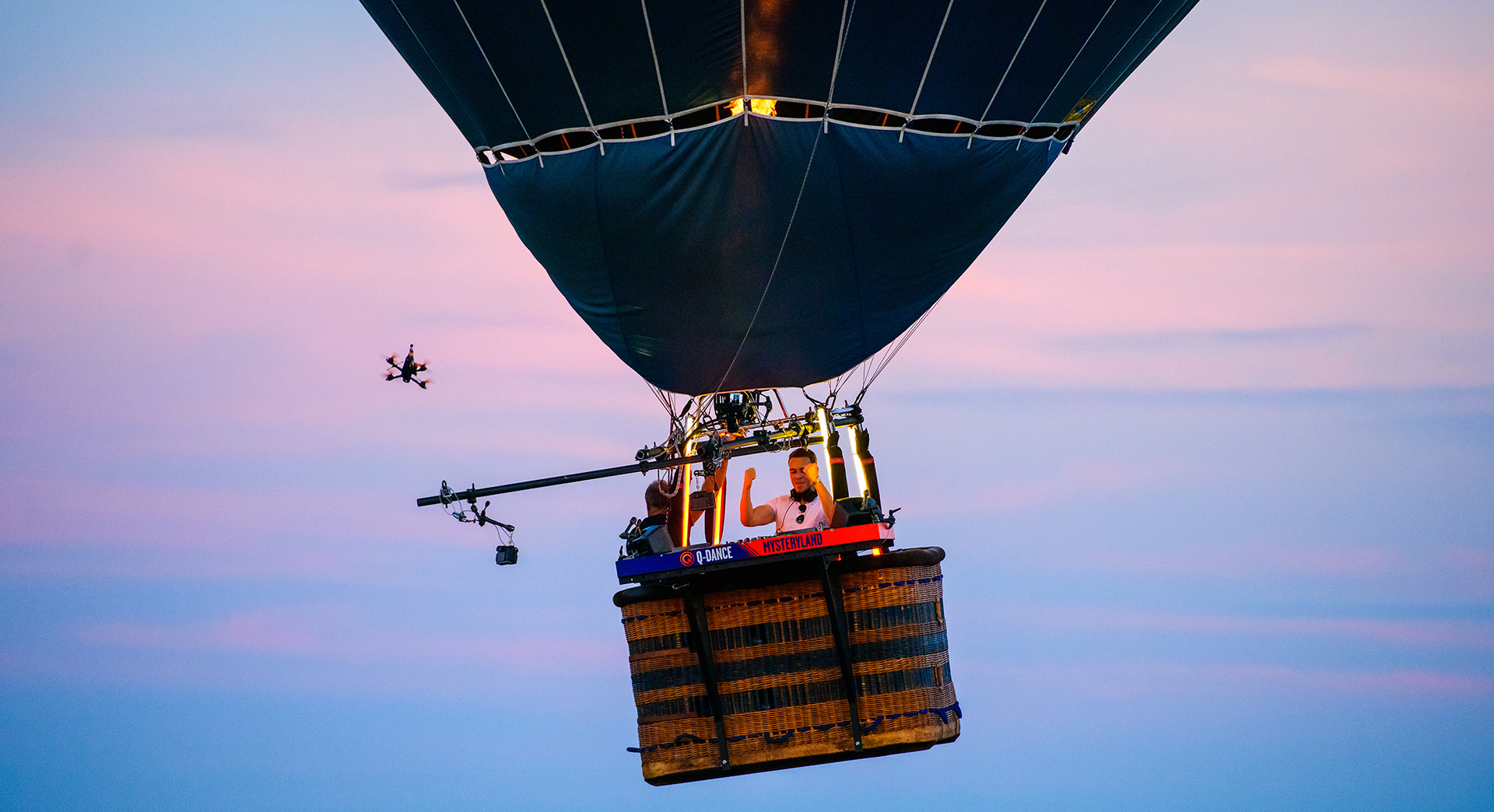 Hot air balloon in flight with a dj playing inside and a drone nearby against twilight sky.