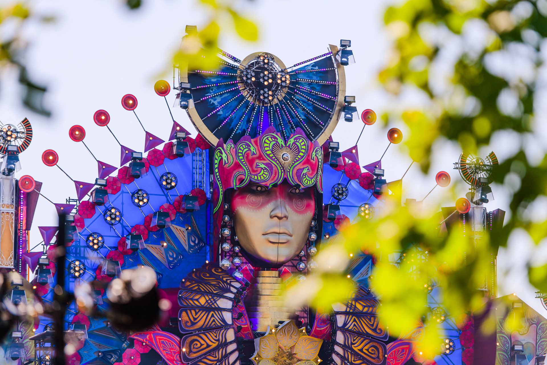 Colorful festival stage with lights and decorations, viewed through foreground leaves.