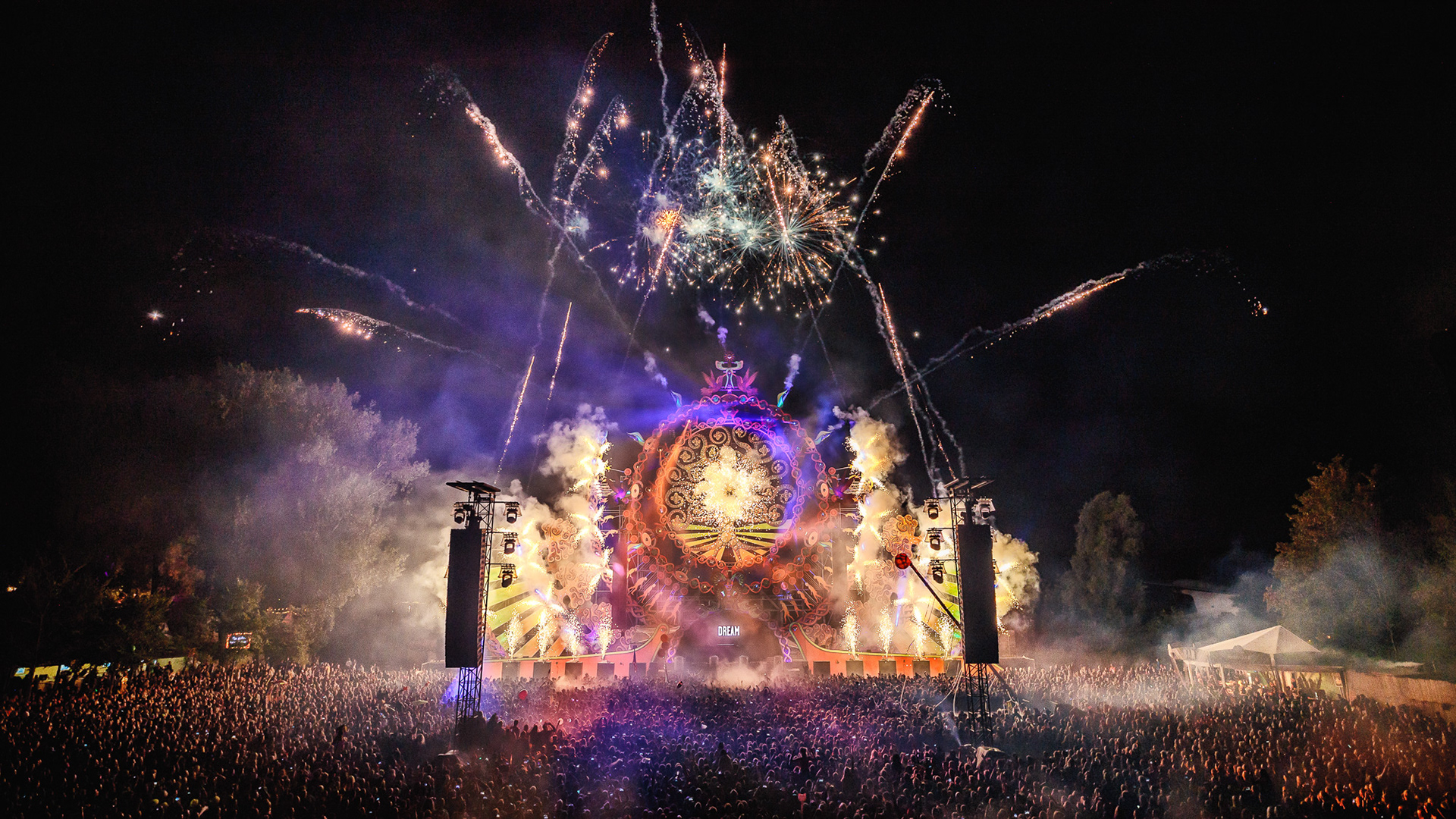 Fireworks burst above a stage with a large crowd at a night festival.