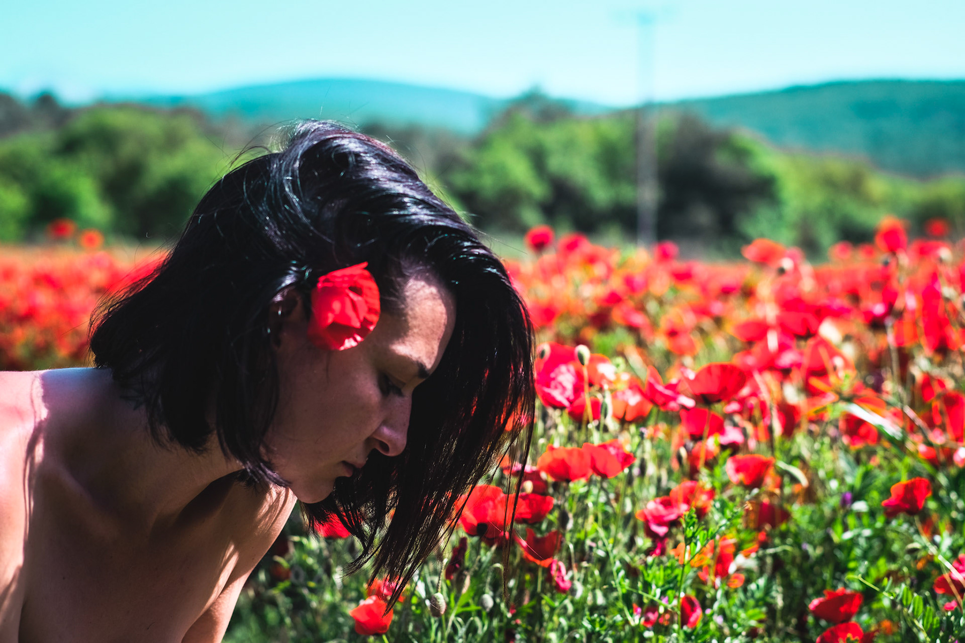 The portrait of a woman with a red flower on her ear. She is leaning over other red poppies.