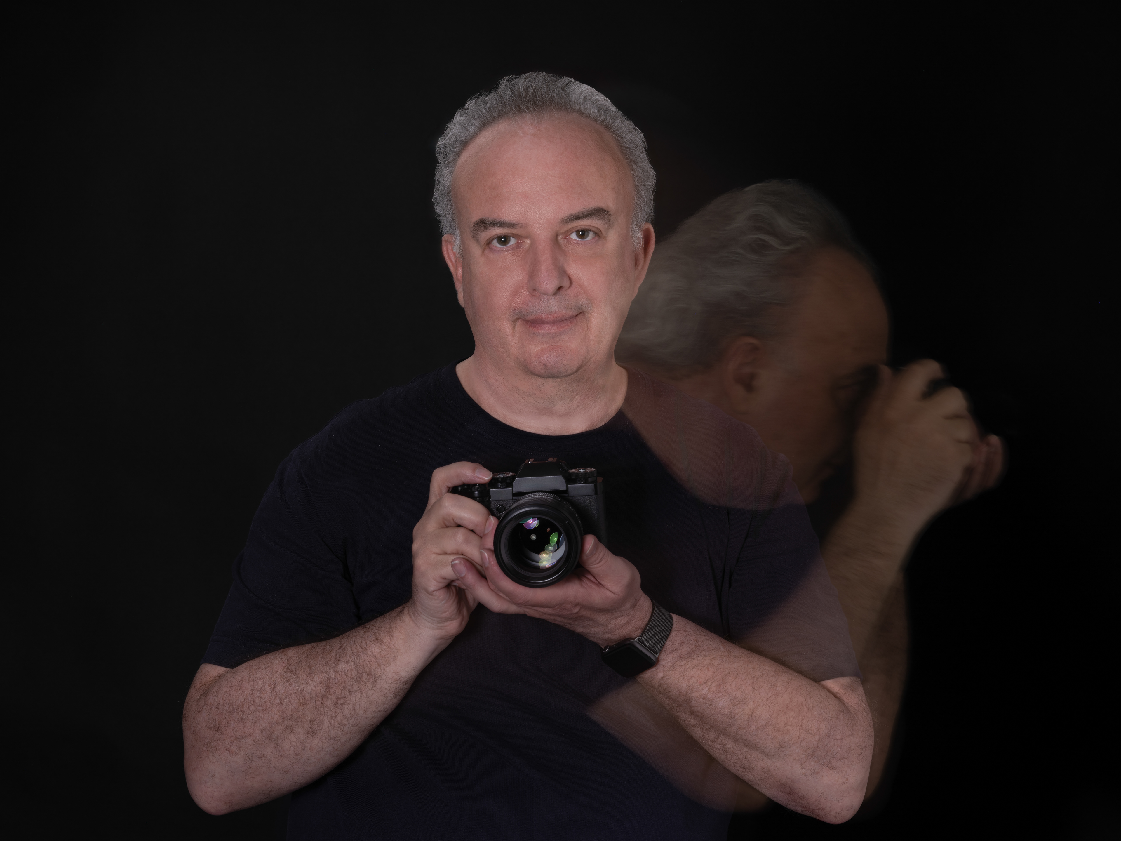 Portrait of photographer Luc Holper holding a camera, with a blurred second figure behind him against a black background.