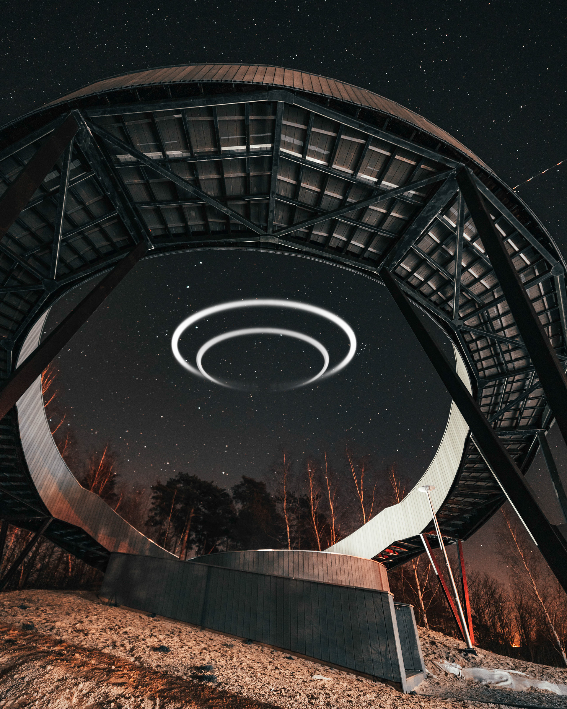 Środek nocy. Taras widokowy w Widuchowej w województwie zachodniopomorskim. Kadr ustawiony jest mniej więcej z pozycji pasa i skierowany ku górze ukazując konstrukcję tarasu, który zbudowany jest na planie okręgu zawieszonego w przestrzeni. W oddali widać las, a środek okręgu wypełniony jest gwiazdami oraz dwoma białymi kręgami świetlnymi unoszącymi się w powietrzu podkreślającymi zjawiskowość tego miejsca. Z obrazu bije zarówno spokój jak i niepewność. Czy jesteśmy sami we wszechświecie?