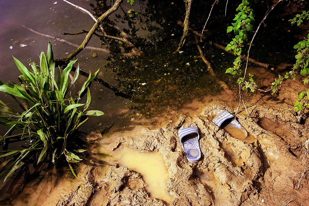 A pair of flip-flops trapped in the mud in front of the stagnant water. Something strange has happened here.