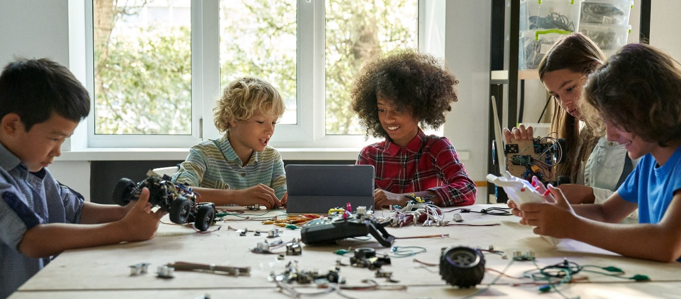 Five kids of different races are sitting at a table, designing an electronic toy car. The boy on the left is holding the car's base. The boy to his right is looking with the black girl at a tablet. The girl is holding a part of the car that has wires on it and the boy on the right is playing with a controller. There are car parts all over the table. Behind the students is a three-paneled window. You can see it is daytime and there are trees outside. On the right of the window are shelves.