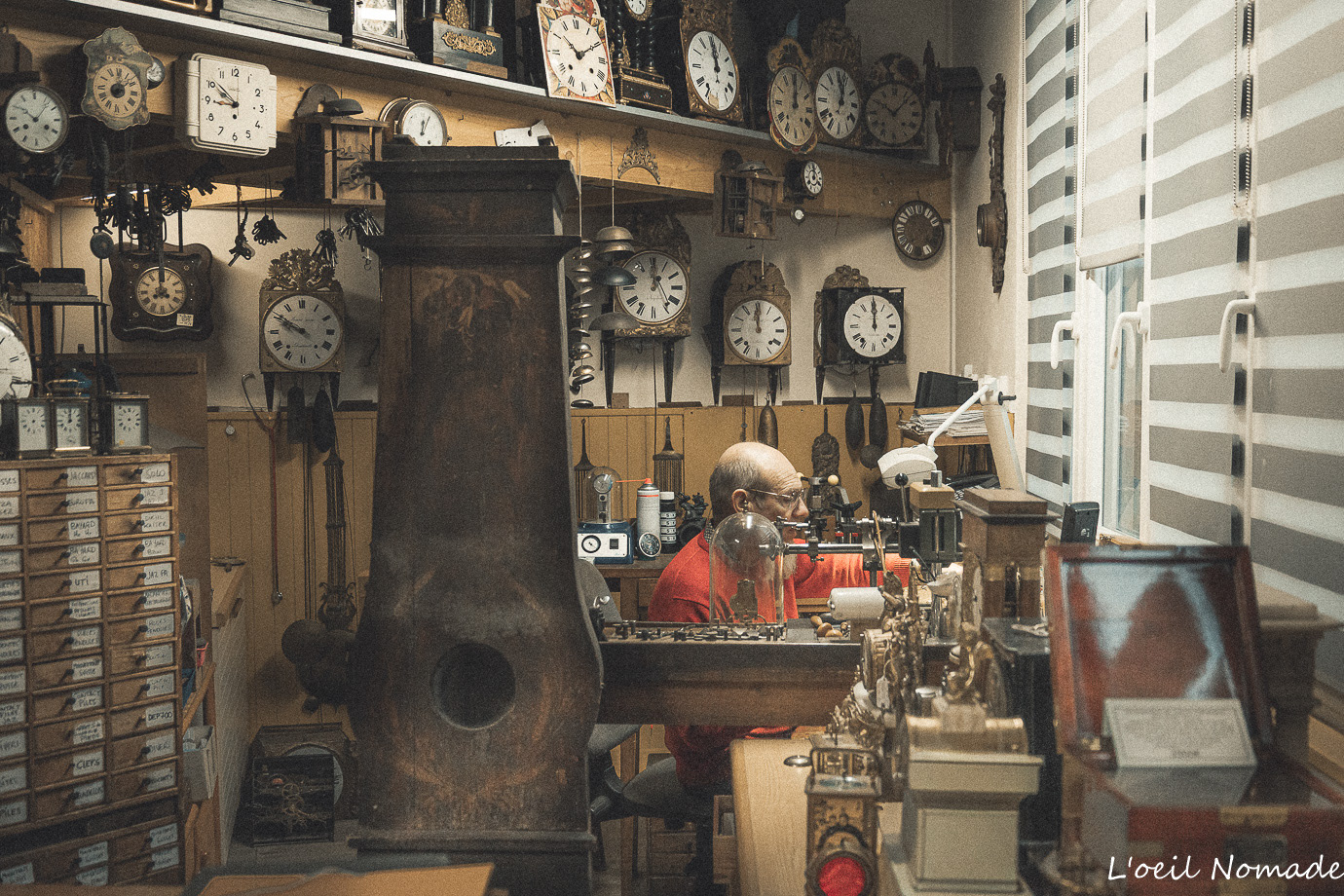 Établi d'horloger encombré d'outils et de pièces anciennes, étagères de fournitures, atmosphère de travail artisanale.