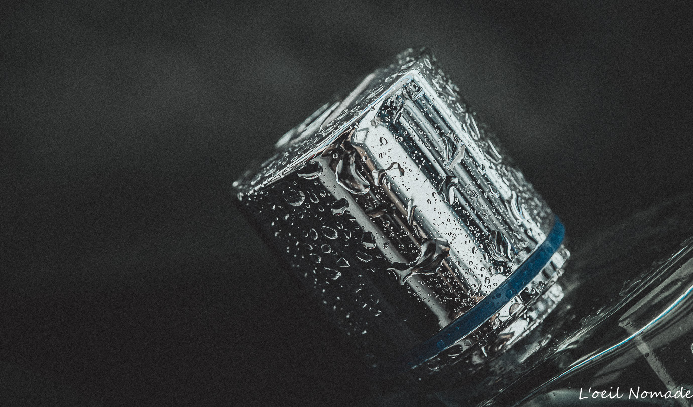 Macro gouttes d'eau sur bouchon de parfum chromé, précision technique et textures minérales.
