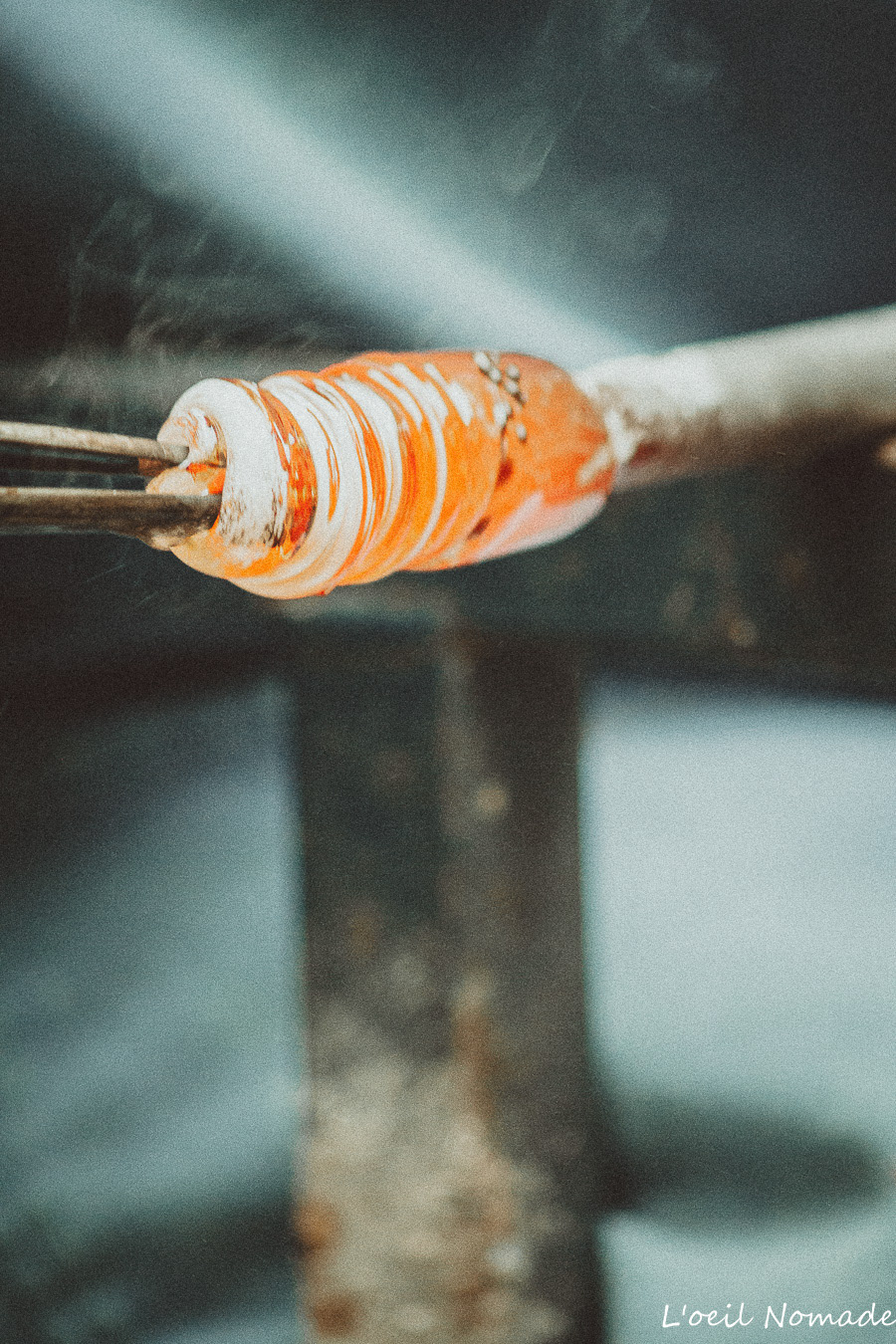 Gros plan sur le travail du verre en fusion : utilisation d'une pince de précision et émanation de fumée, photographie documentaire révélant la beauté brute et l'exigence du métier.
