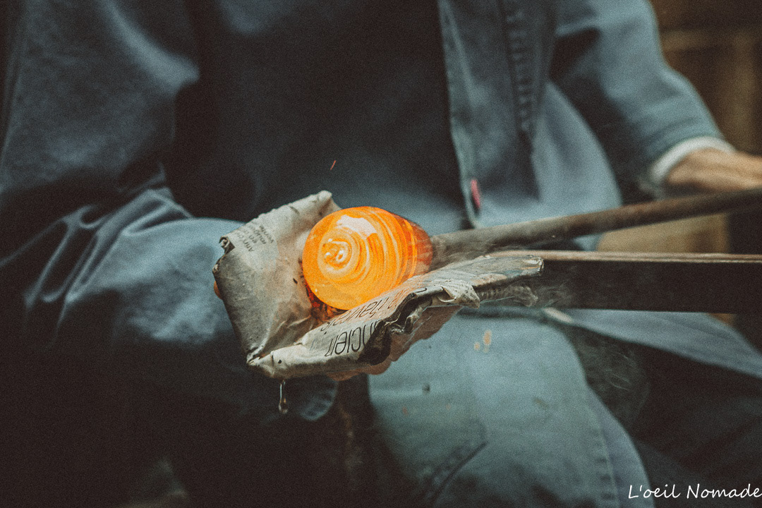Photographie d'artisanat d'art : geste technique d'une souffleuse de verre façonnant la matière en fusion avec du papier journal humide, focus sur l'interaction entre les éléments.