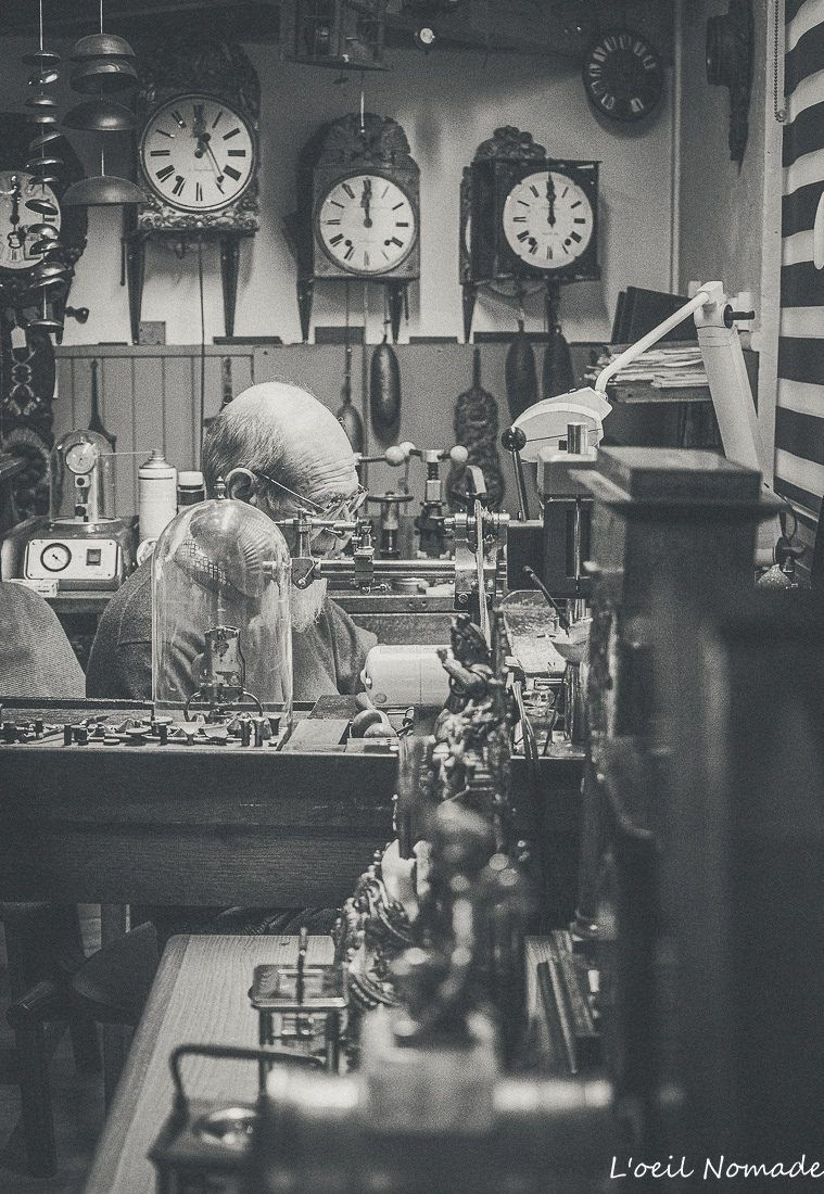 Atelier d'horlogerie avec horloges comtoises au mur, ambiance authentique et intemporelle en noir et blanc.