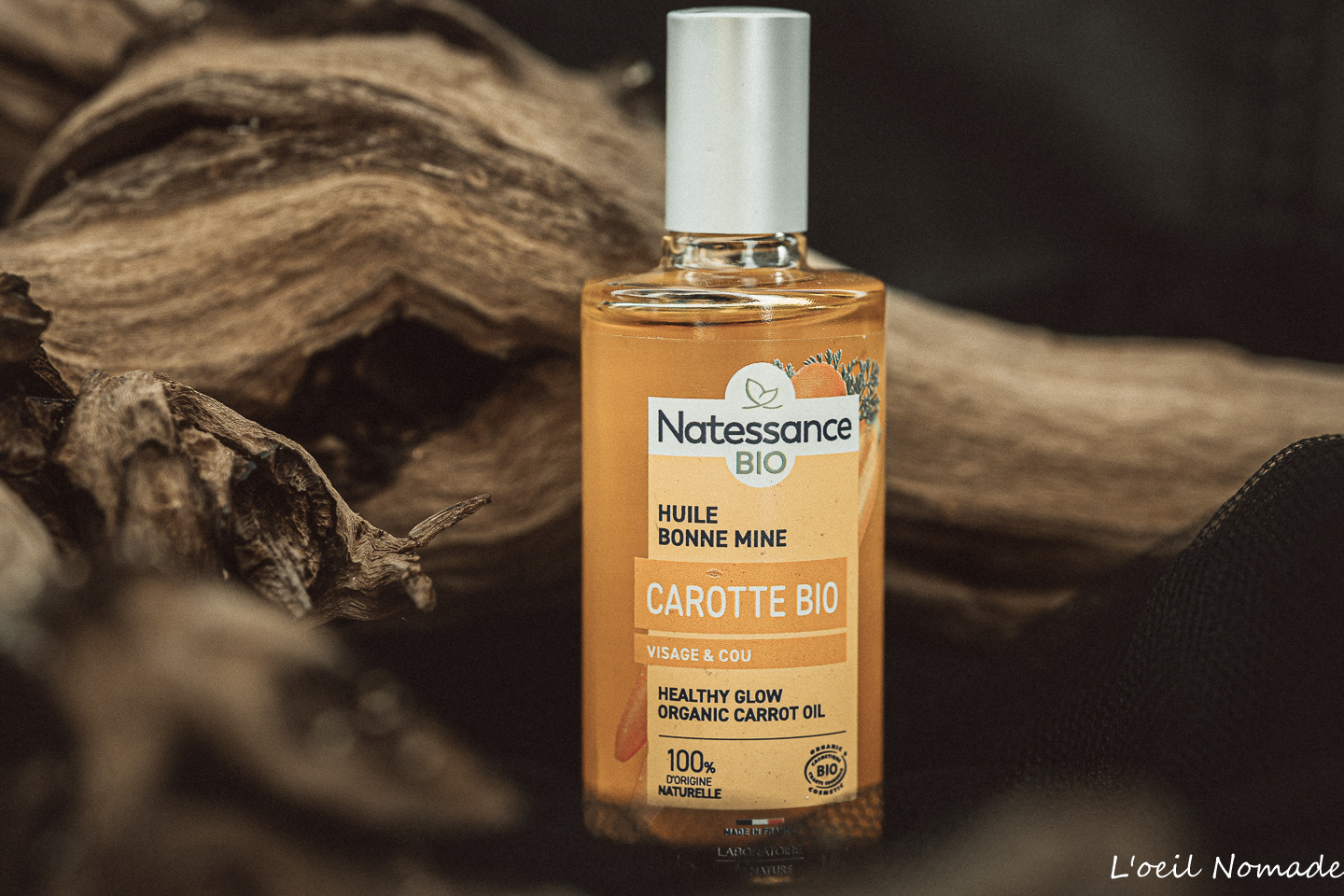 Bouteille d'huile de carotte cosmétique premium, mise en scène avec bois flottés et texture naturelle, éclairage contrôlé, L'œil Nomade.
