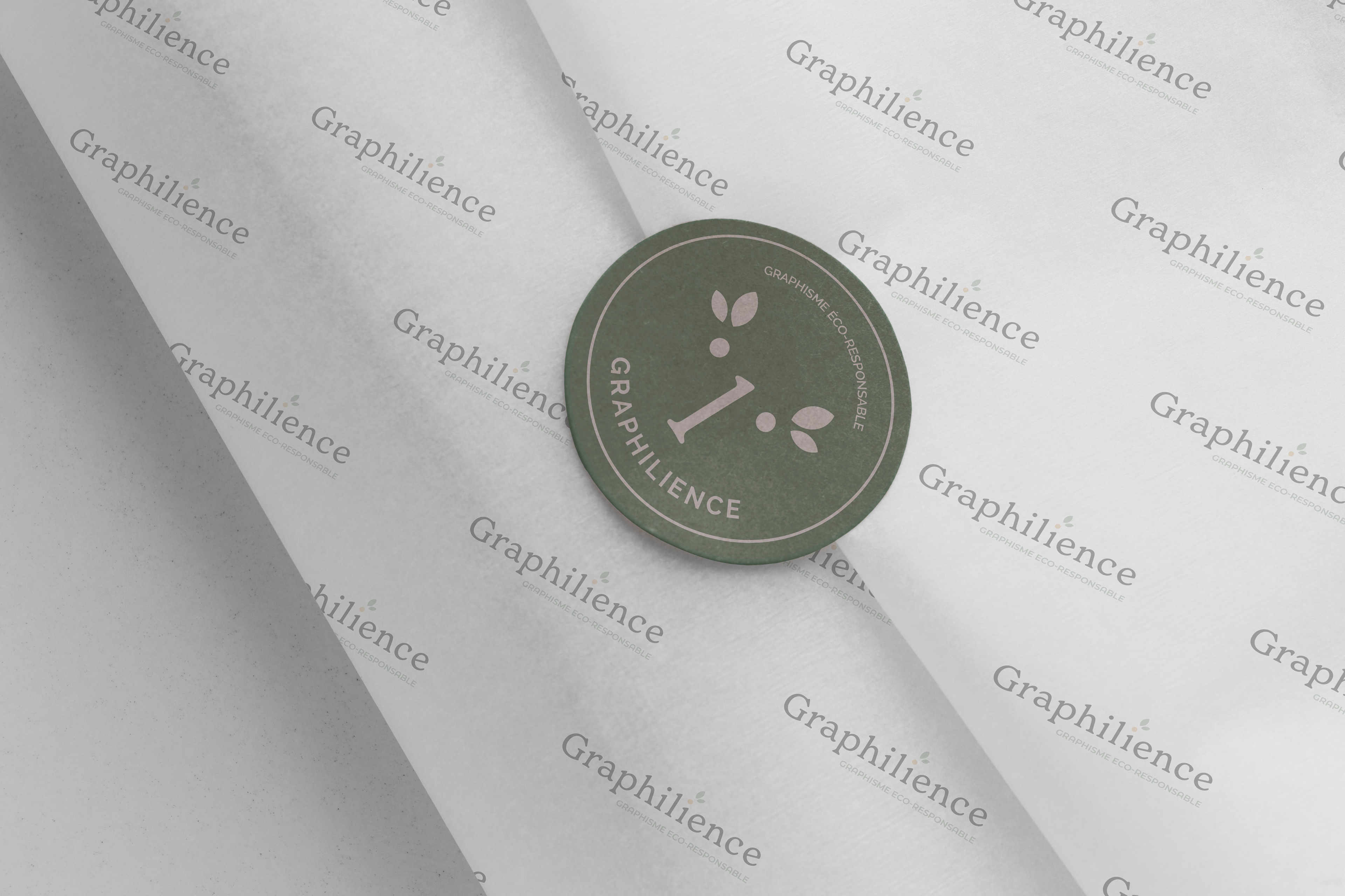 Papier de soie imprimé avec le logo Graphilience et un autocollant vert portant le pictogramme de Graphilience, représentant un visage souriant avec des feuilles stylisées et le texte 'GRAPHILIGENCE - GRAPHISME ECO-RESPONSABLE'.