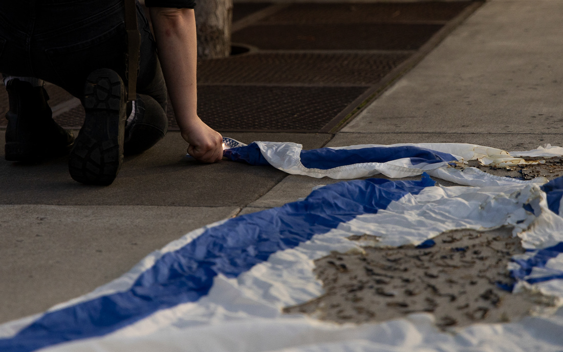 A demonstrator attempts to reignite the partially-burnt Israeli flag outside of the EMU as the march continues on Oct. 7, 2024. Protesters gathered for the anniversary of the Hamas-led attacks on Israel to protest the ongoing Israel-Hamas war and show solidarity with its victims. The march began at the Wayne Lyman Morse U.S. Courthouse and ended at the corner of E 18th Ave. and University St. | Shot for Emerald Media Group | 1/60 f/4 ISO 1600