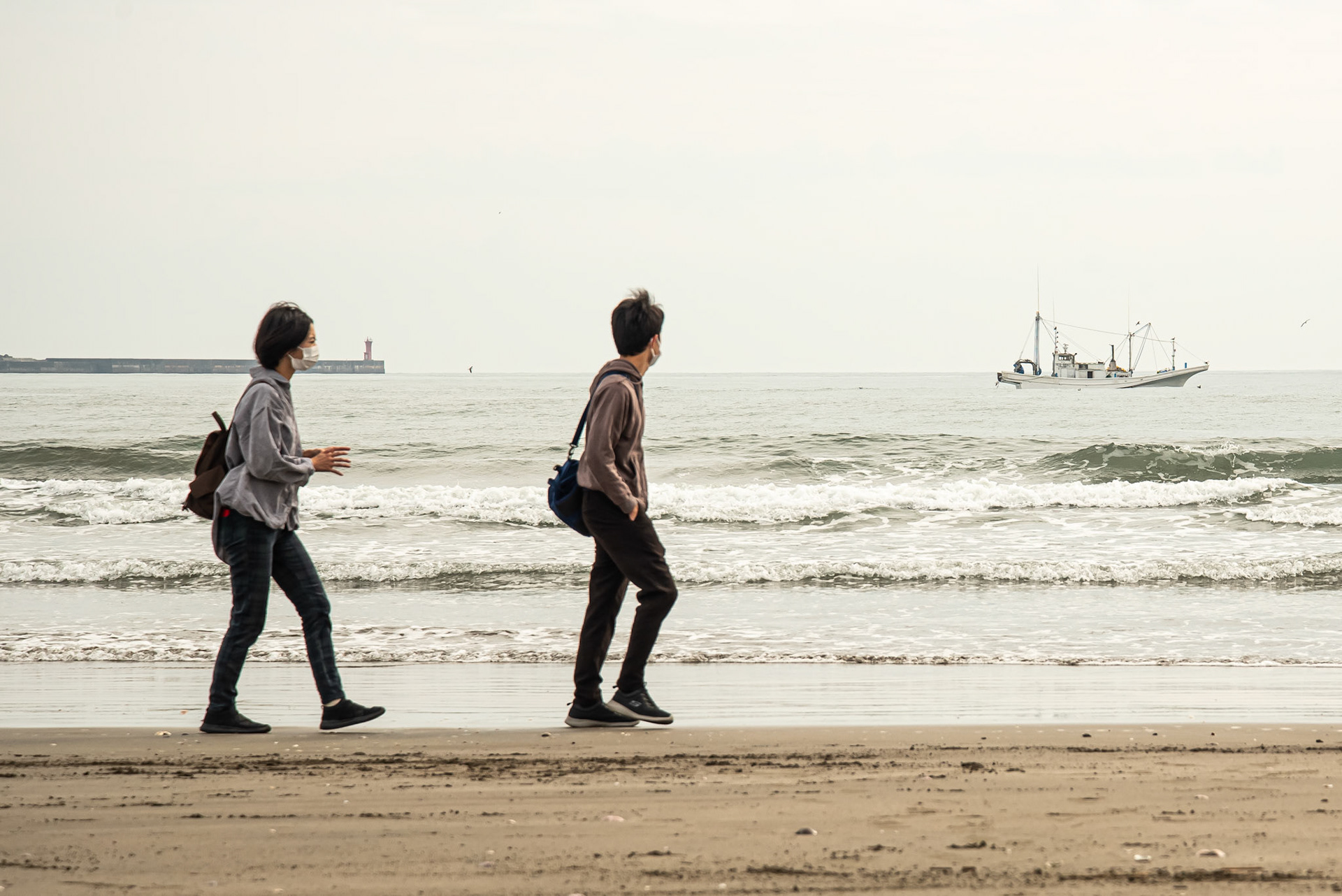 Un couple marche le long de l'eau sur la plage au Japon avec en arrière fond la mer les vagues un bateau et une digue sous un ciel gris