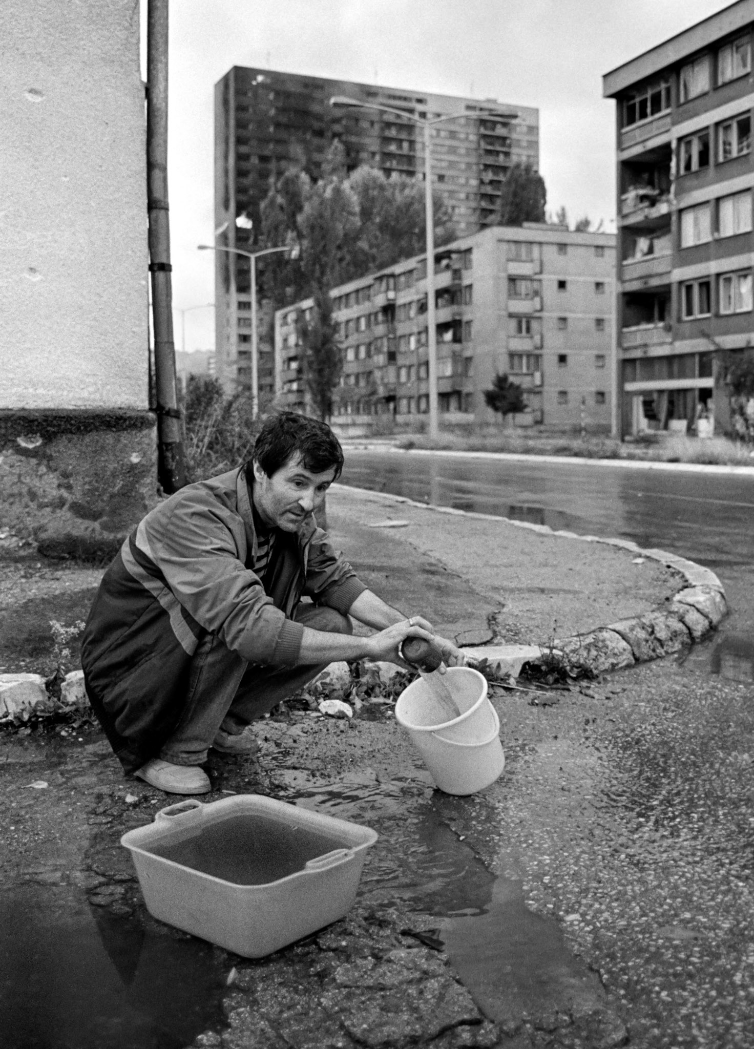 A man gathers rainwater from a puddle in the street in the besieged Bosnian capital Sarajevo on November 28 1993, while a block of flats burns from artillery shelling in the background. 350,000 Sarajevans have lived mostly without water, electricity and gas since April 1992.
