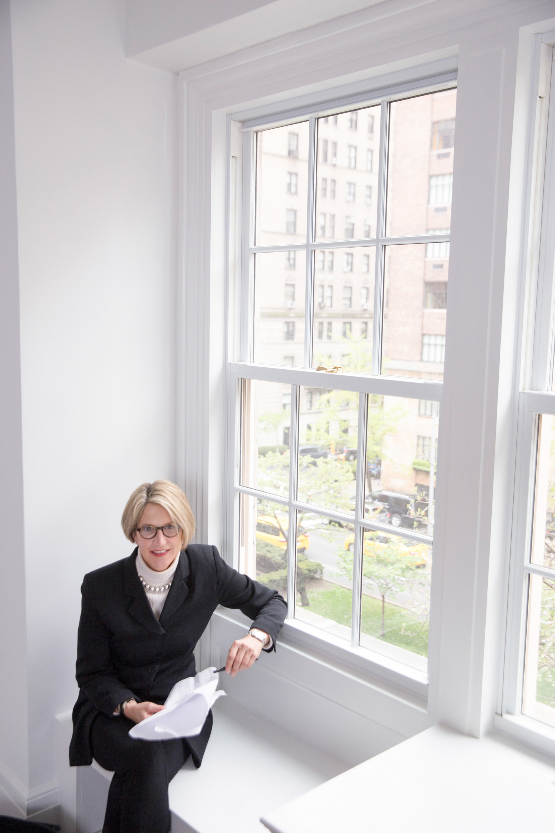 Lifestyle portrait of NYC luxury realtor studying architectural floor plans with city skyline visible through window.