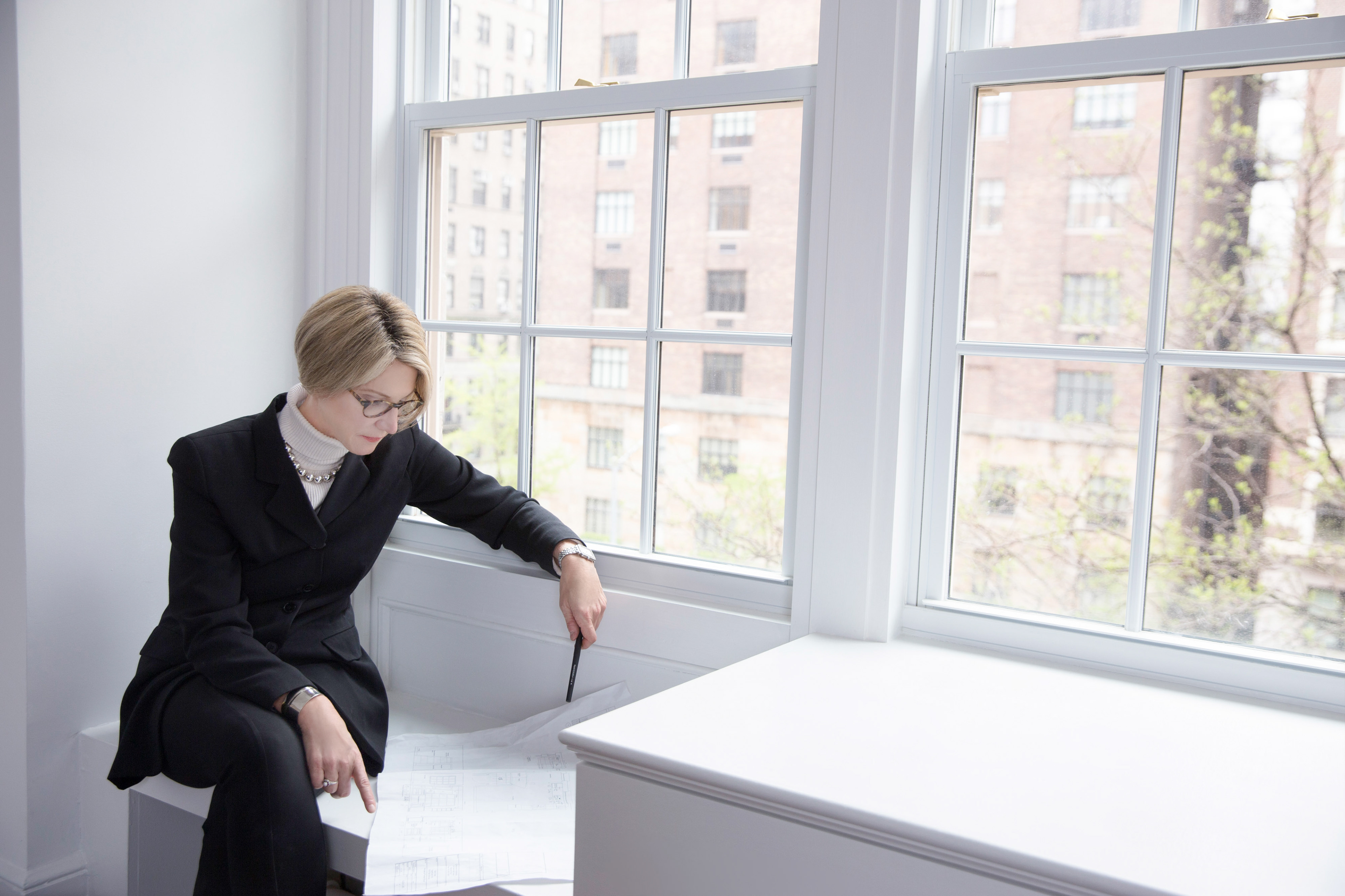Environmental portrait of New York City luxury real estate agent reviewing apartment blueprints in front of large Manhattan window.