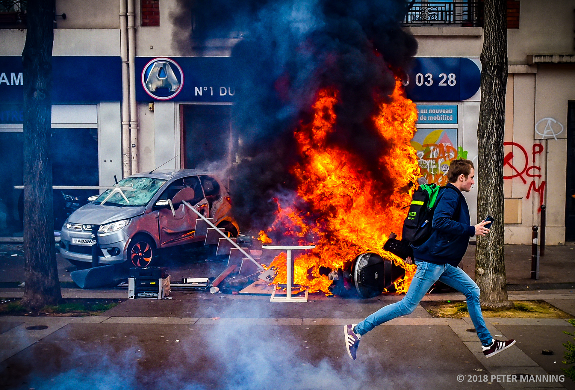 A young man runs past a burning motorbike after 'Black Bloc' protesters smashed their way into a Renault showroom and caused extensive damage during a May Day march in Paris, France on Tuesday 1st May 2018..