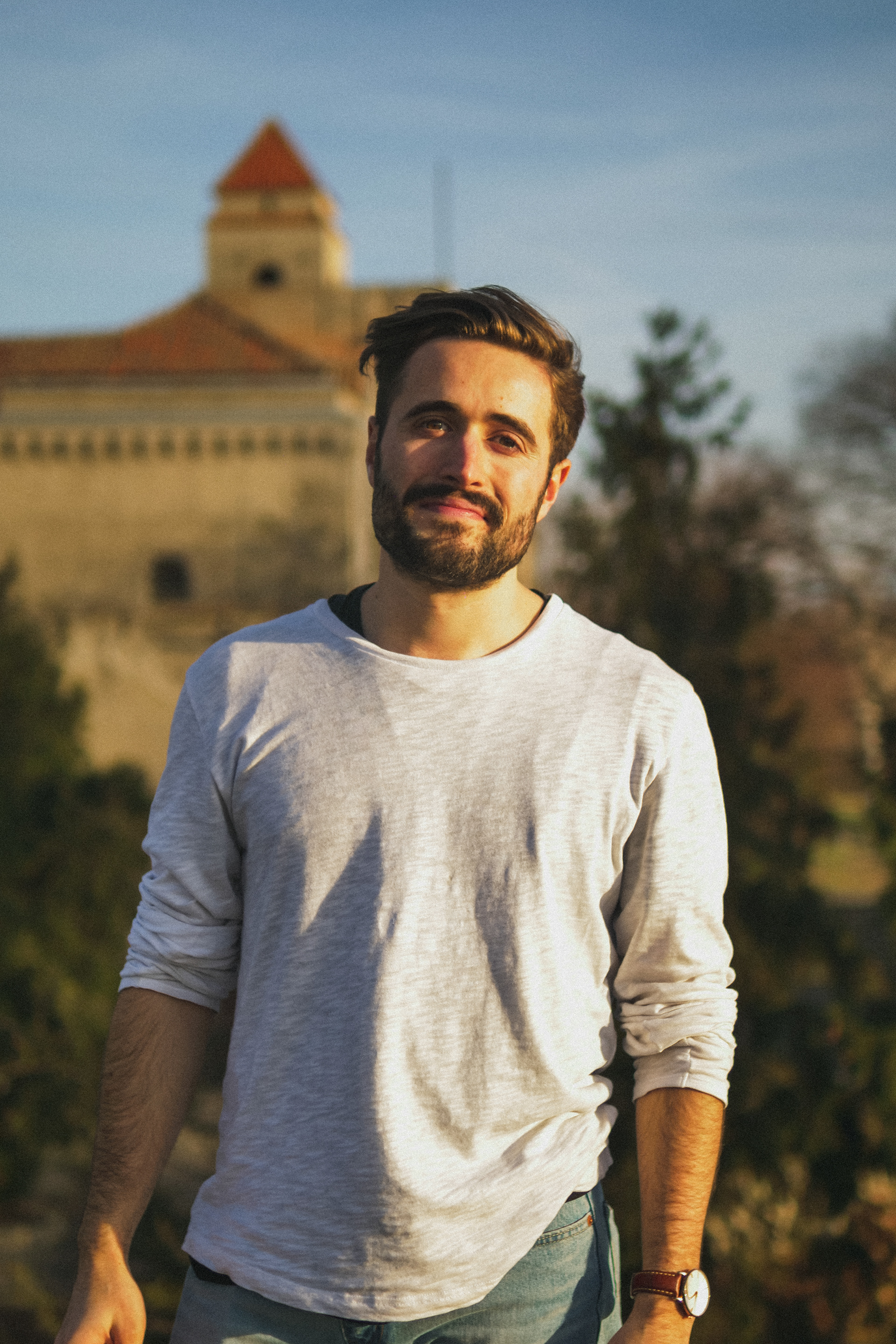 Andrew John standing in front of a castle smiling wearing a white long sleeve top at sunset