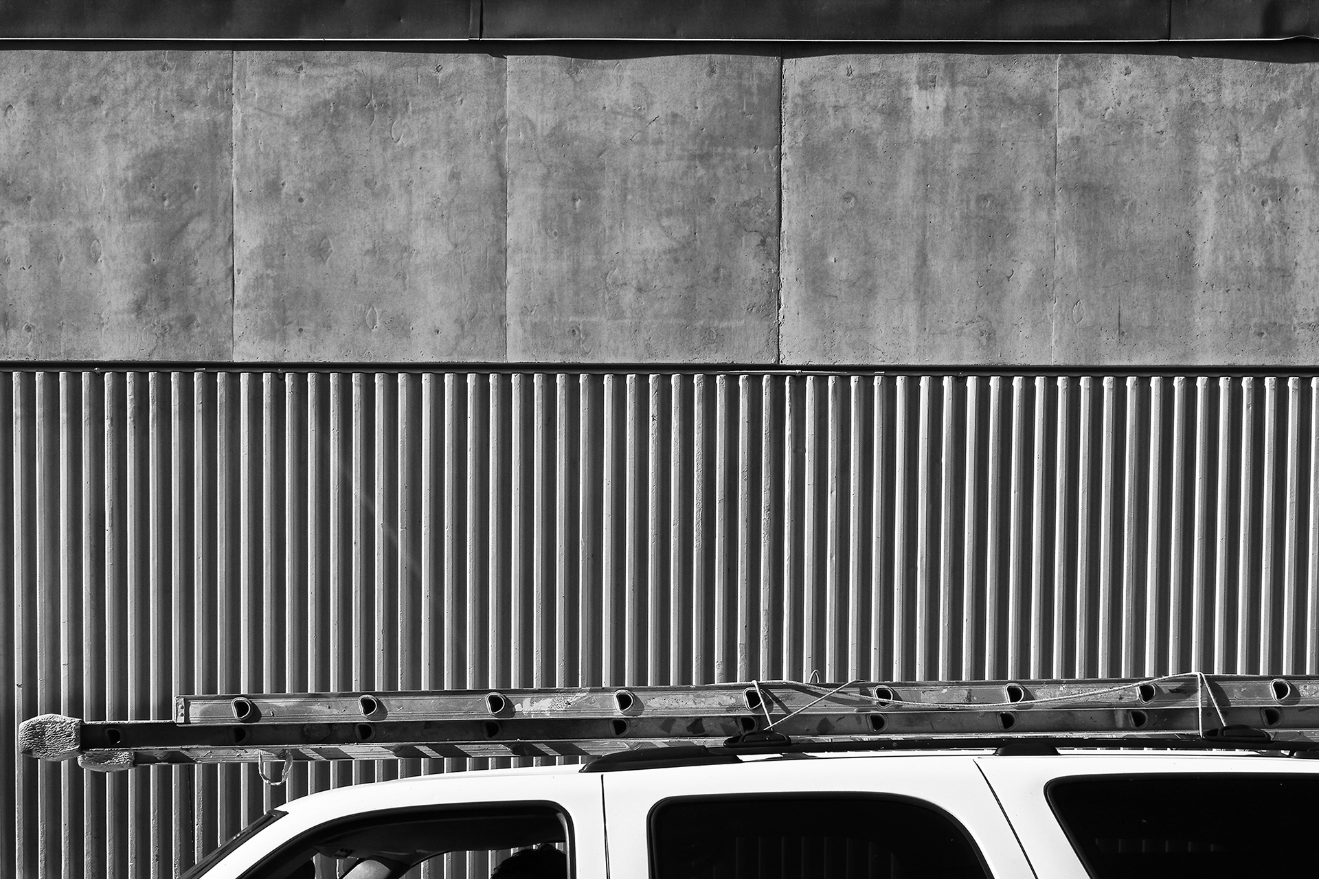 A work truck with a ladder on roof against a stark concrete building, black and white photograph